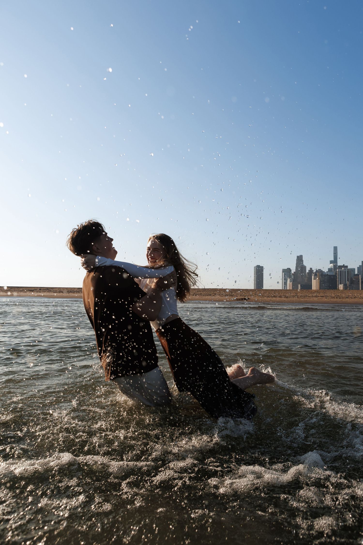 Two people splash and play in the ocean waves with a city skyline visible in the background.