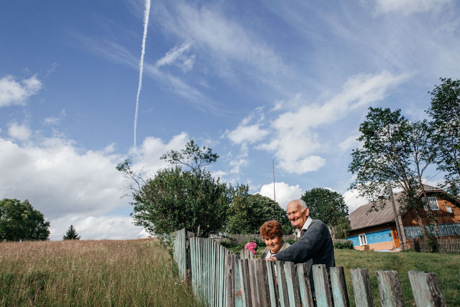 A couple leans against a wooden fence in a meadow under bright blue summer sky with clouds.