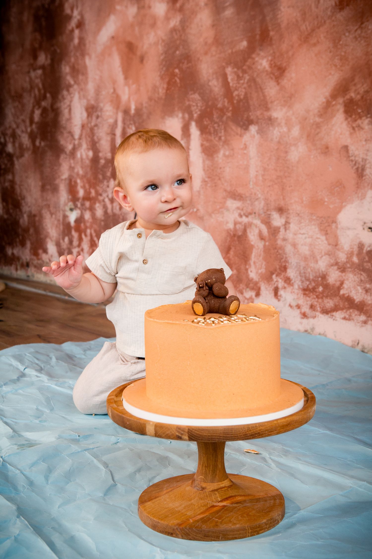 Baby in white outfit sitting next to orange birthday cake with teddy bear topper.