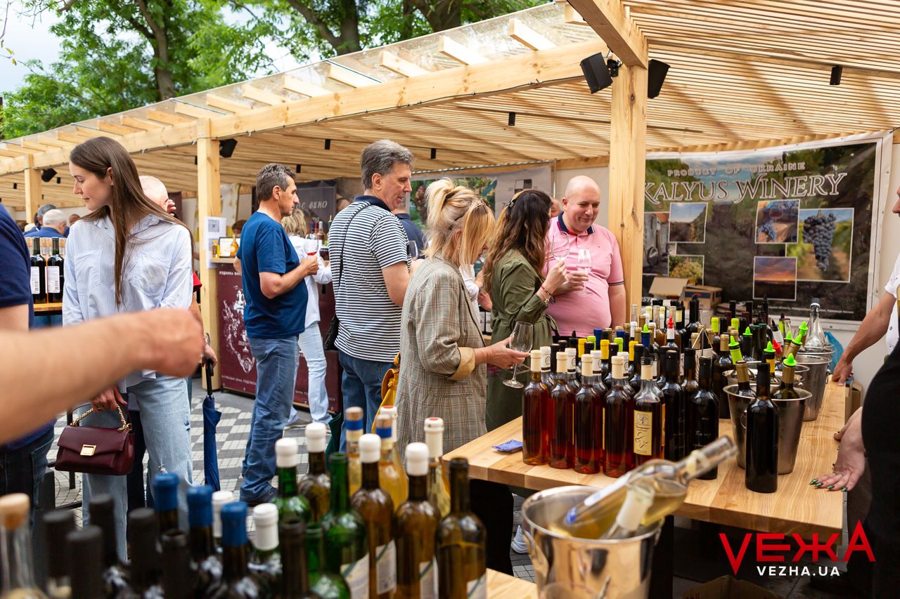 Customers browse wine bottles at an outdoor market under a wooden pergola structure.