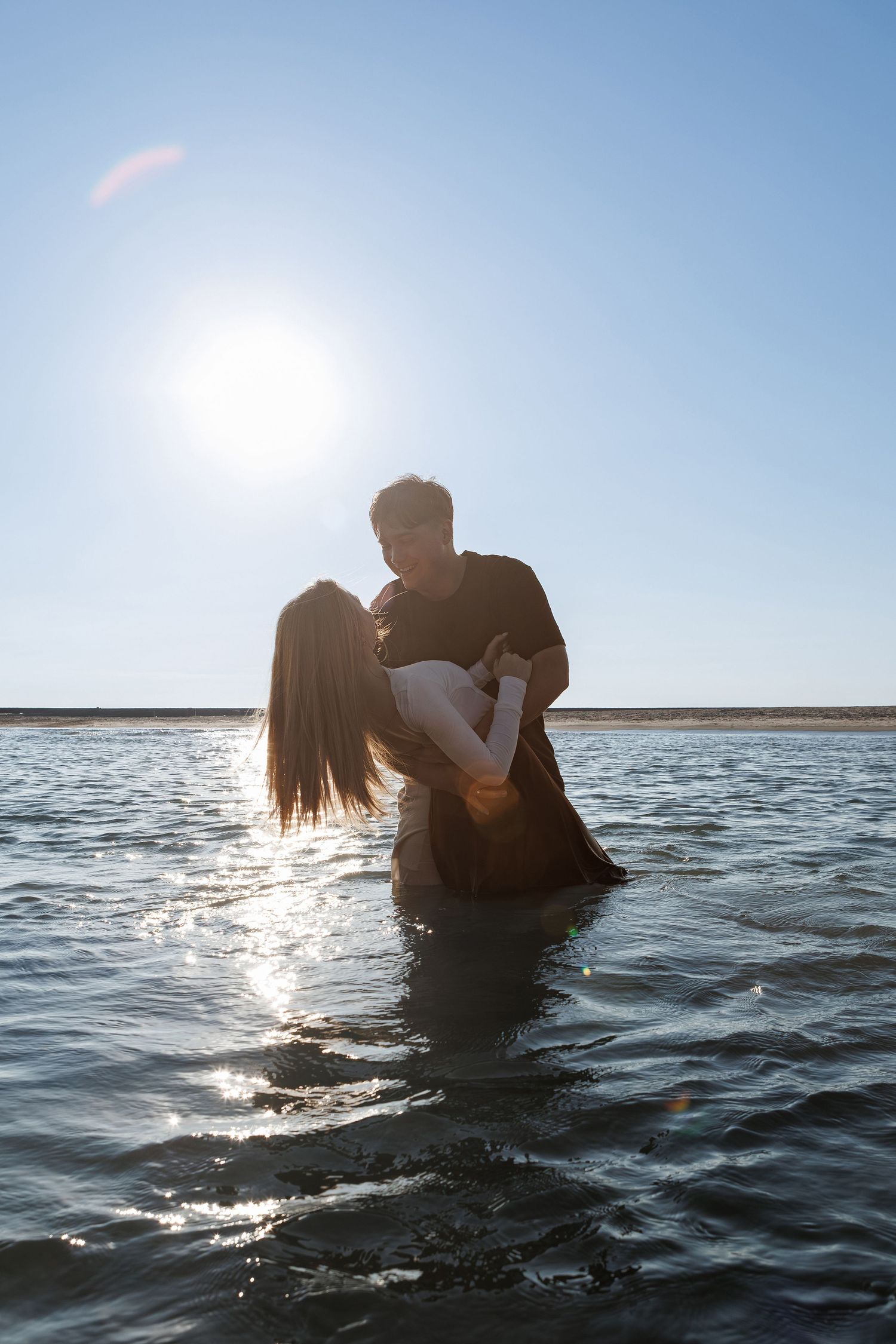 Silhouetted couple embracing in ocean water during sunset with sunlight creating a romantic backlit glow.