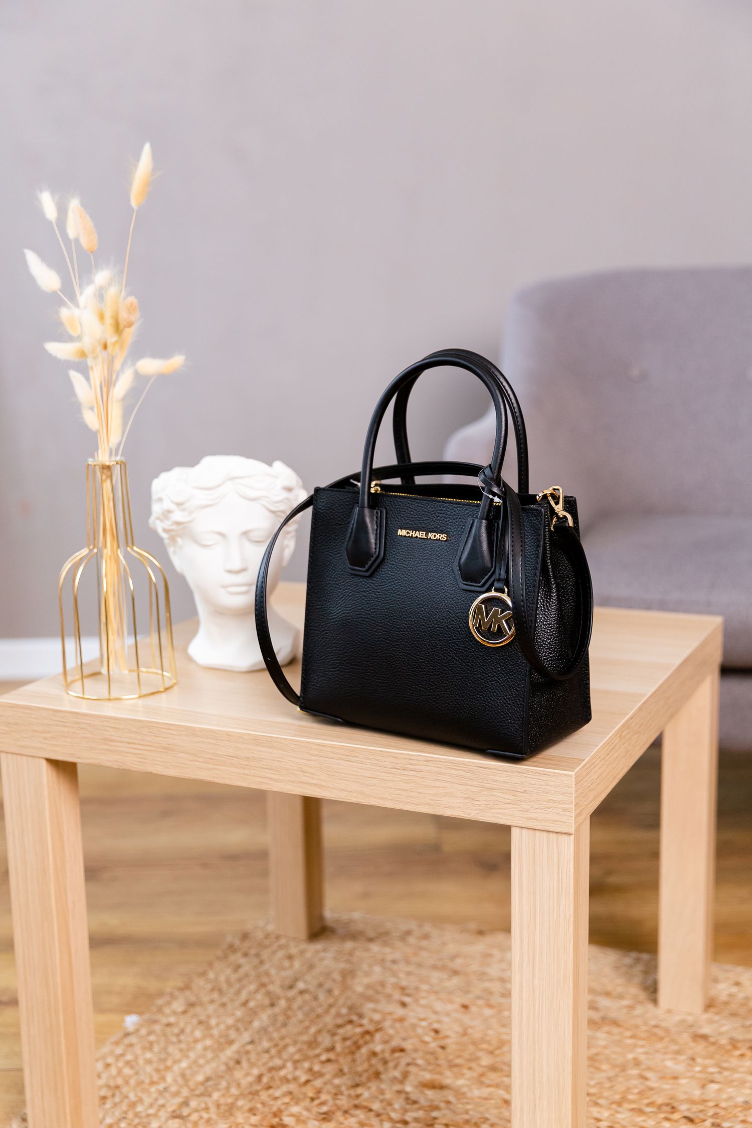 Black leather Michael Kors handbag displayed on wooden table with decorative vase and bust.