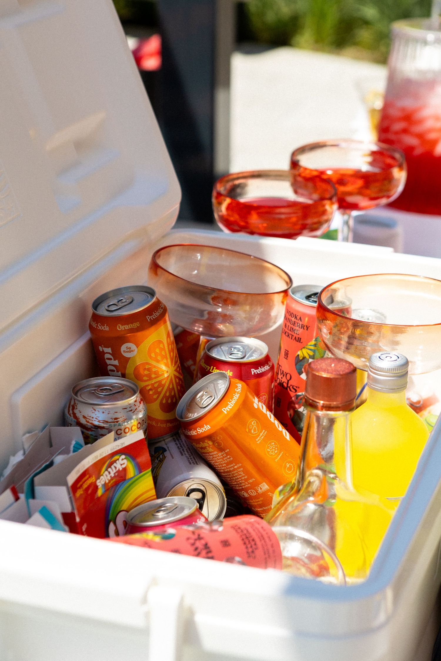 An ice-filled cooler containing colorful canned beverages and drinks with red cocktails in glasses on top.
