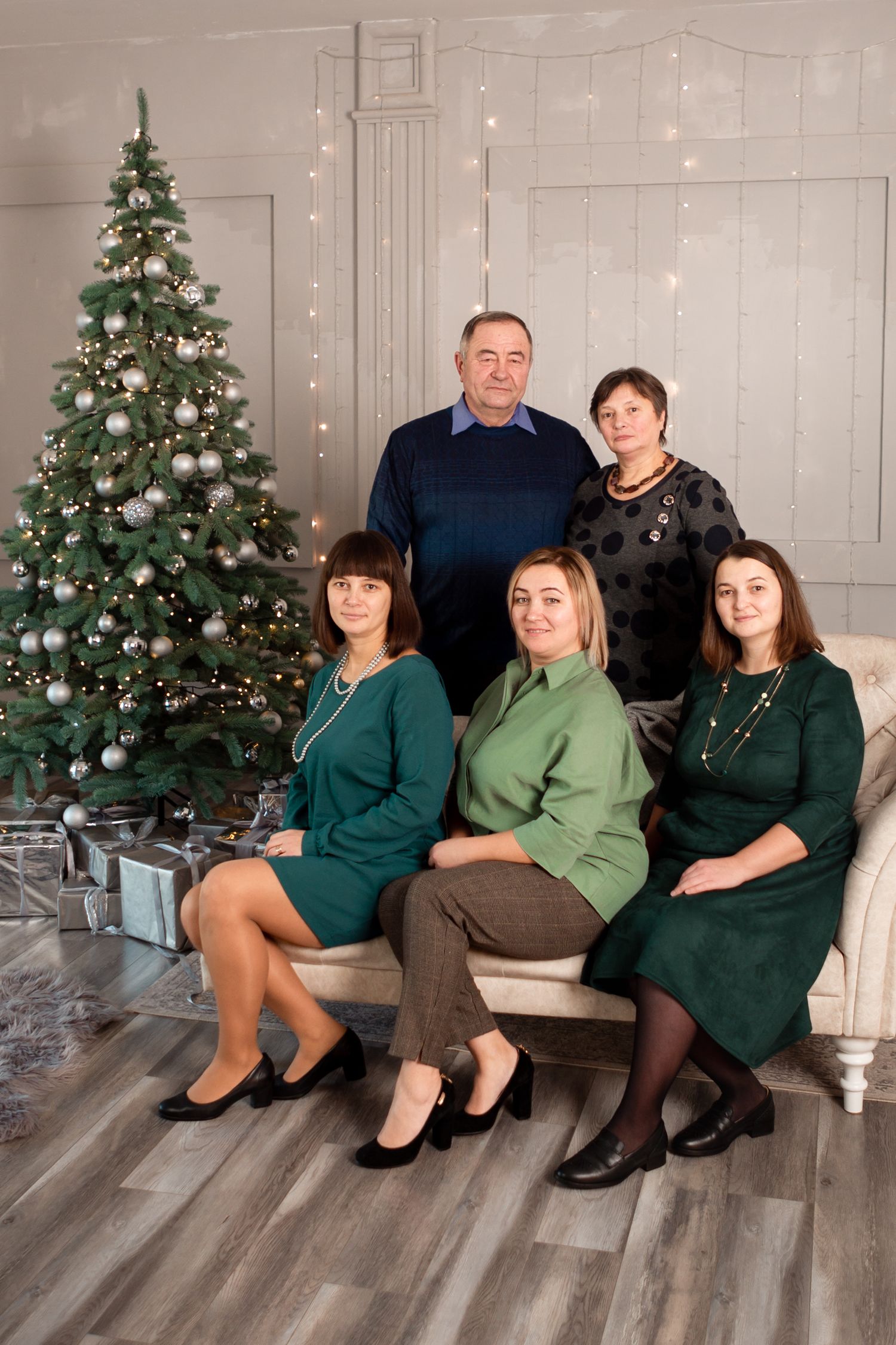 Family group in coordinated green outfits pose formally by Christmas tree with wrapped presents.