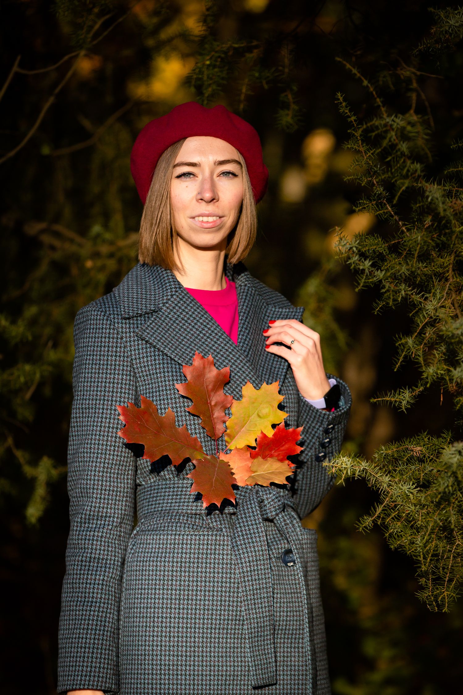 Stylish fall outfit featuring checked coat and burgundy beret while holding autumn leaves.