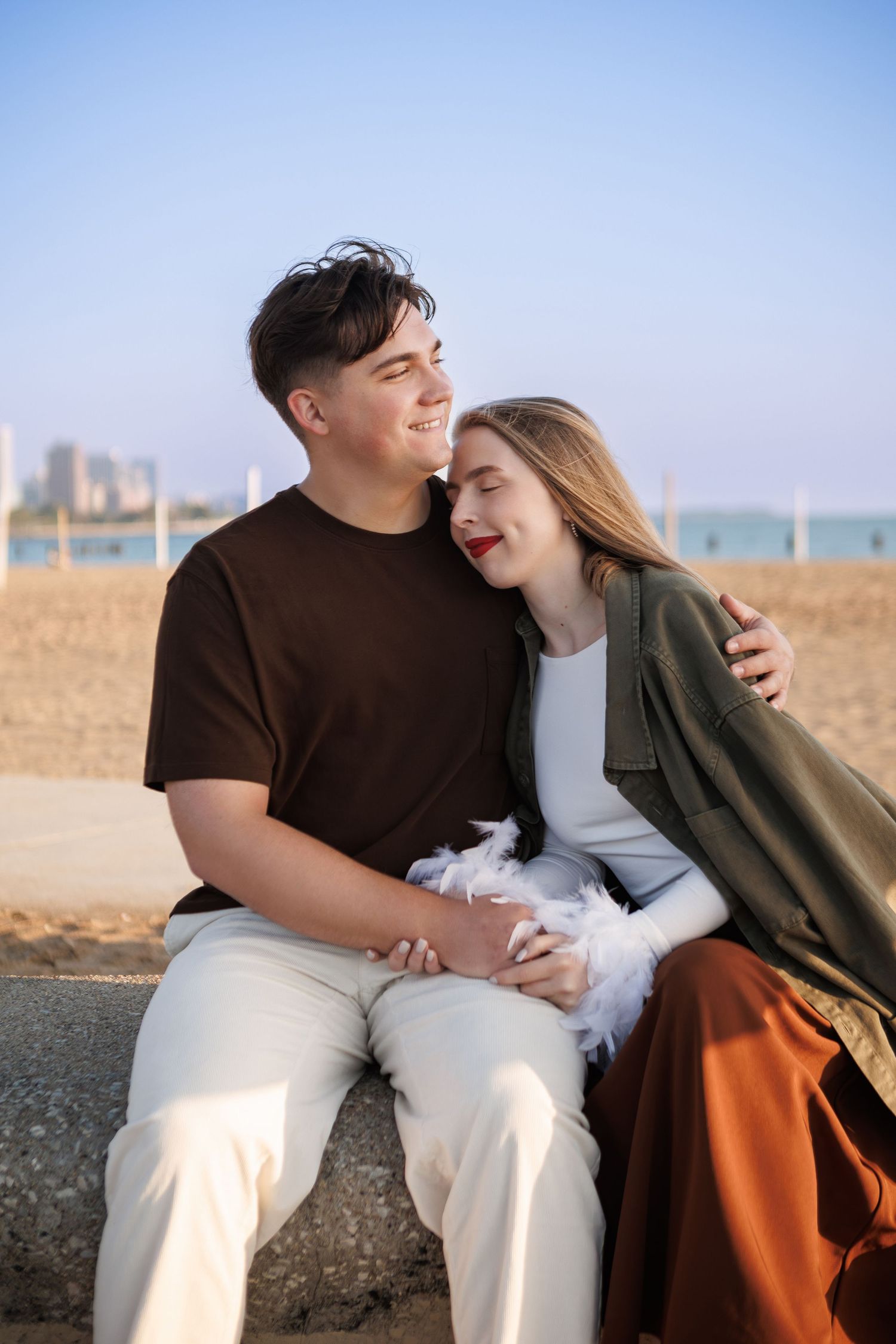 A couple shares a tender moment while sitting together on a concrete barrier at the beach during sunset.