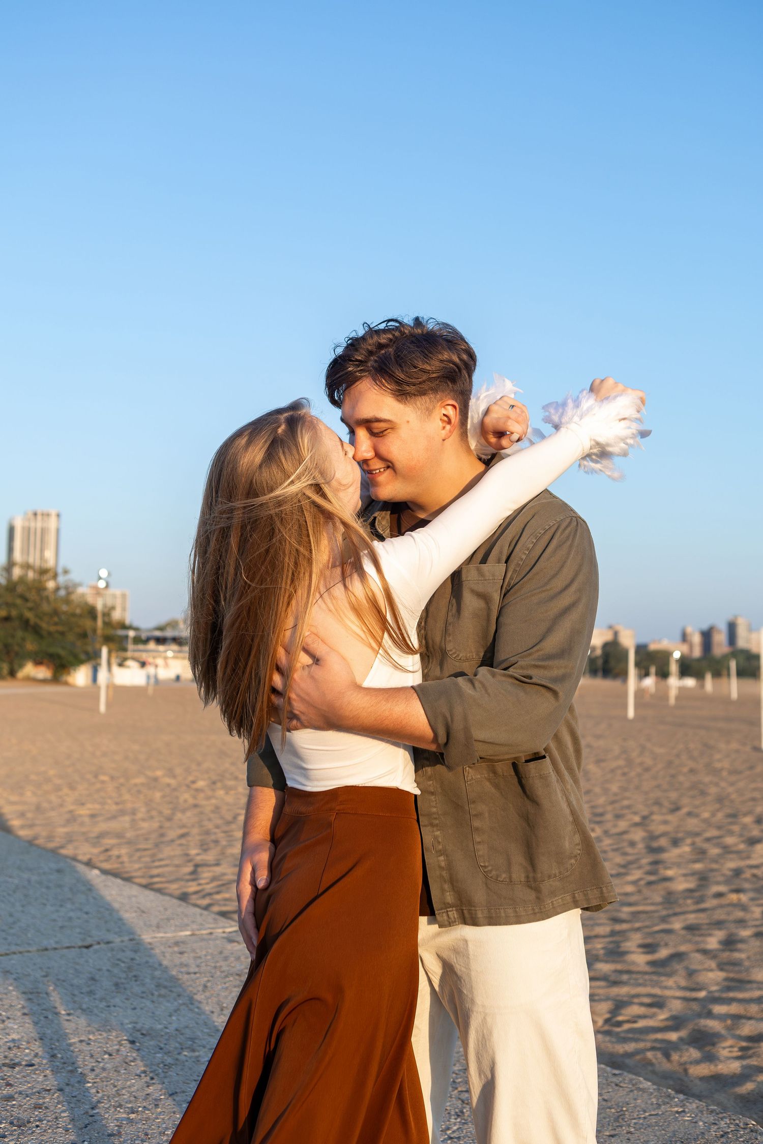 A couple shares a romantic moment on a sandy beach at sunset with buildings visible in the background.