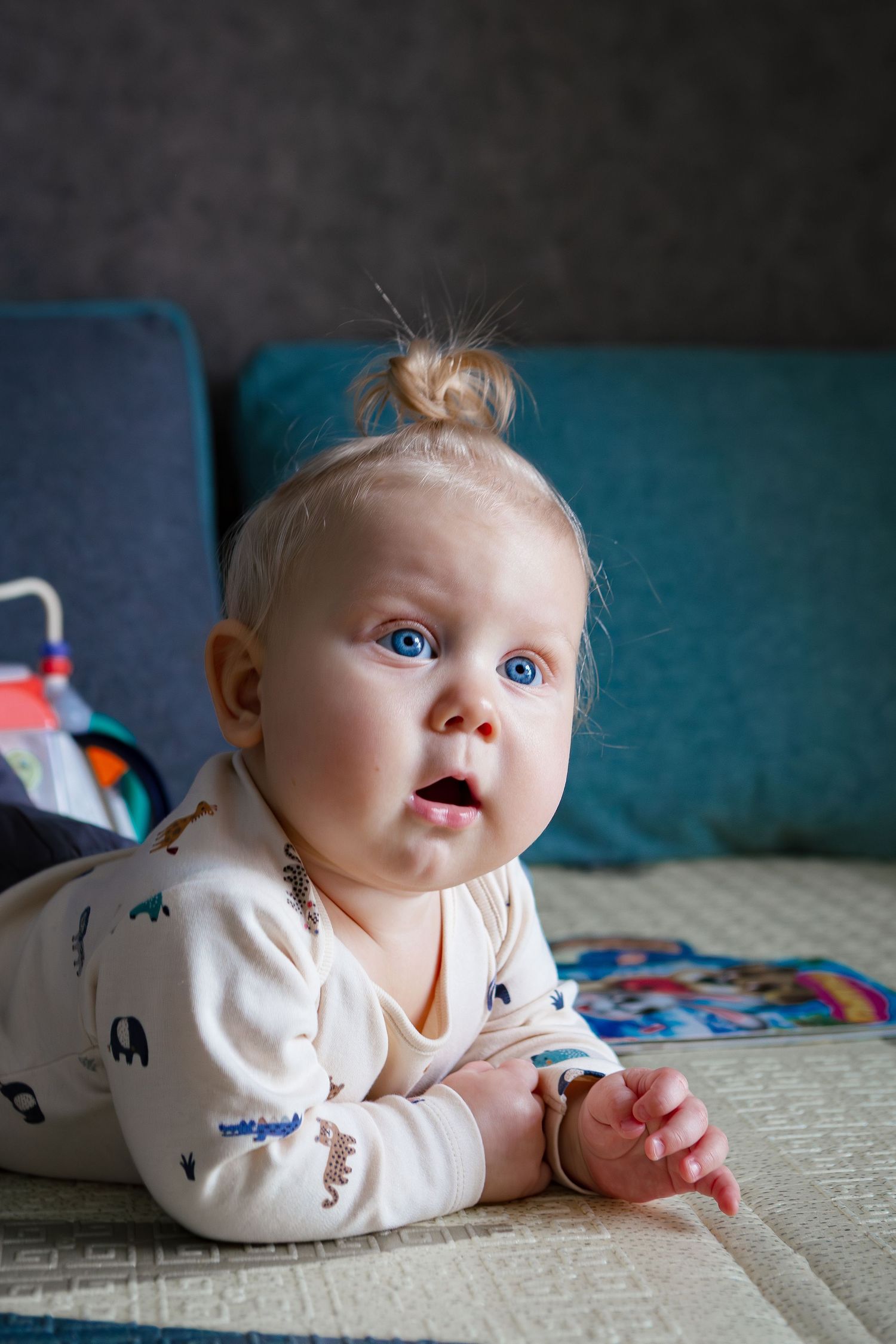 A baby in white patterned sleepwear lies on a couch with striking blue eyes and a tiny top knot hairstyle.