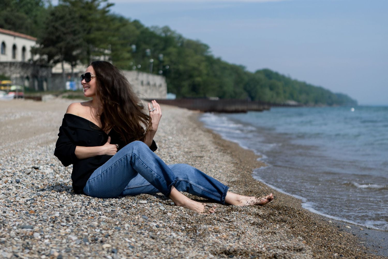 Person rests along a rocky lakeshore with mountains and trees in the background during a sunny day.