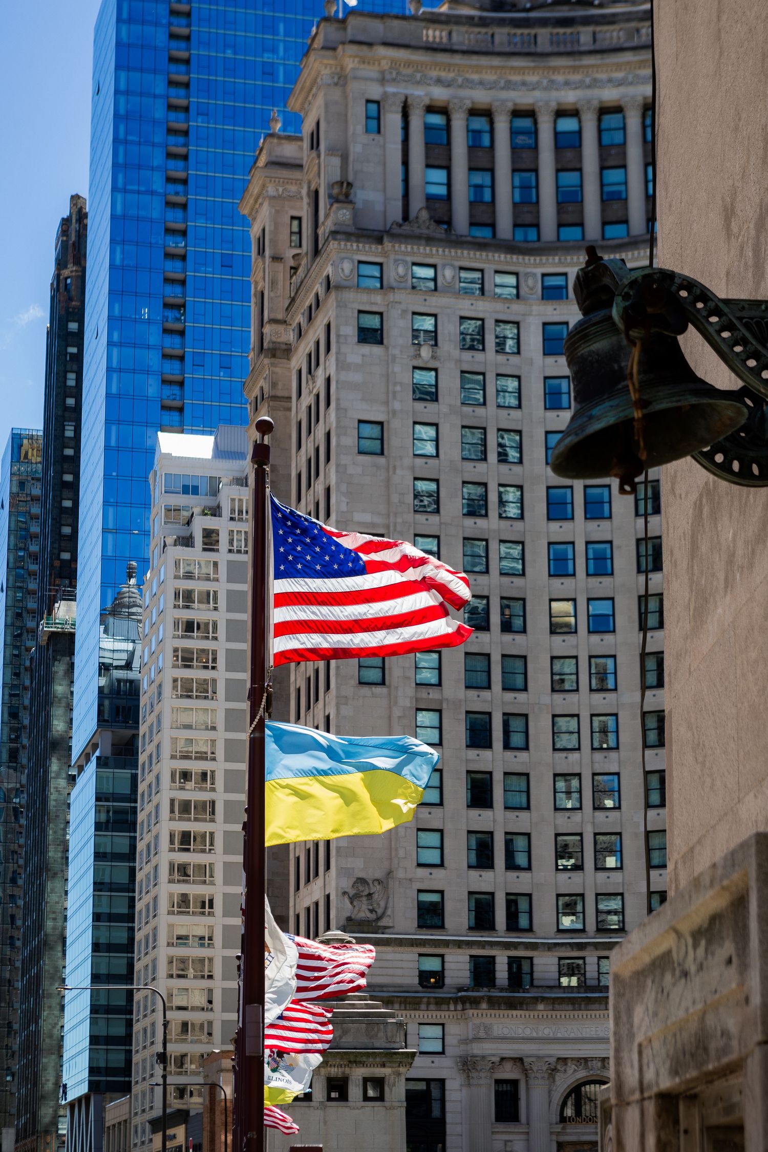 American and Ukrainian flags wave together against backdrop of city buildings on sunny day.