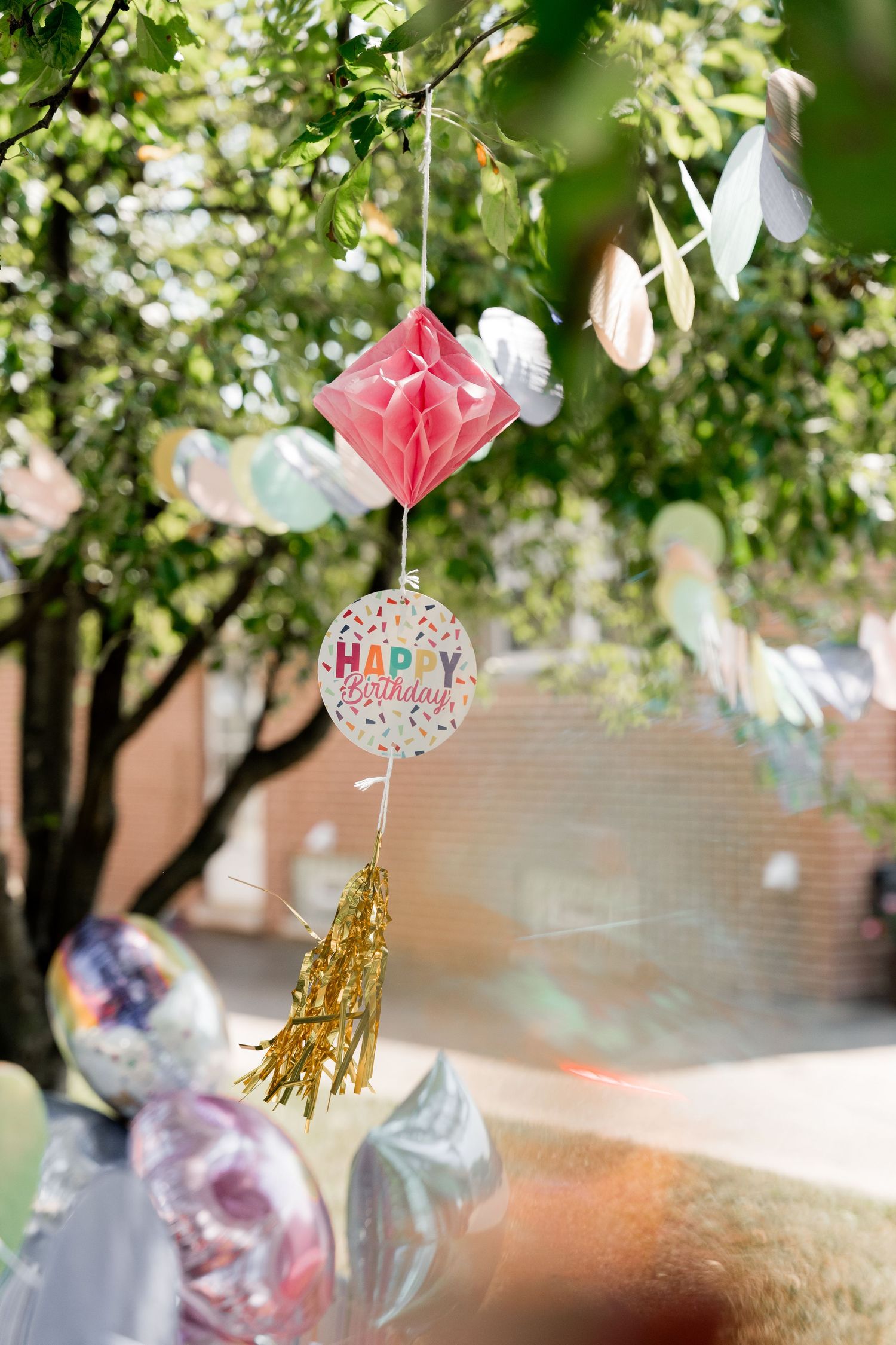 Birthday party decorations with pink paper flowers and gold tassels hanging from tree branches.