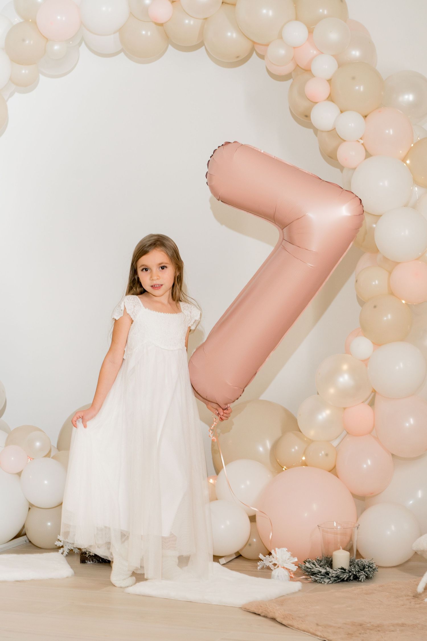 Young girl in white dress posing with large pink number 7 balloon and decorative arch.