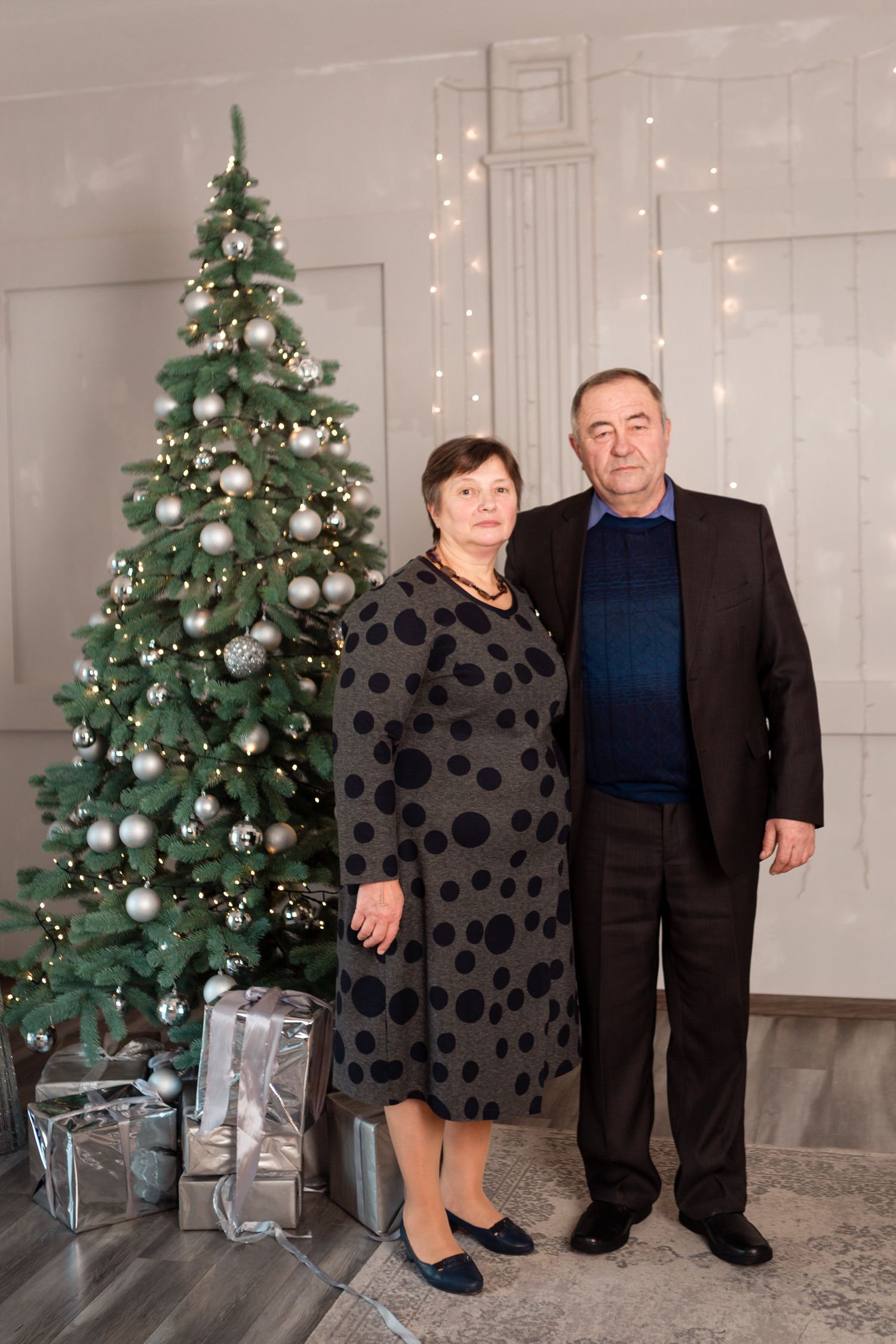 Couple poses together by decorated Christmas tree with silver wrapped presents at base.