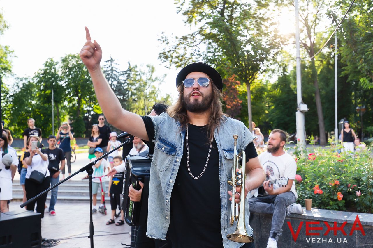 Musician in denim vest and black shirt performing outdoors at a summer concert in a park.