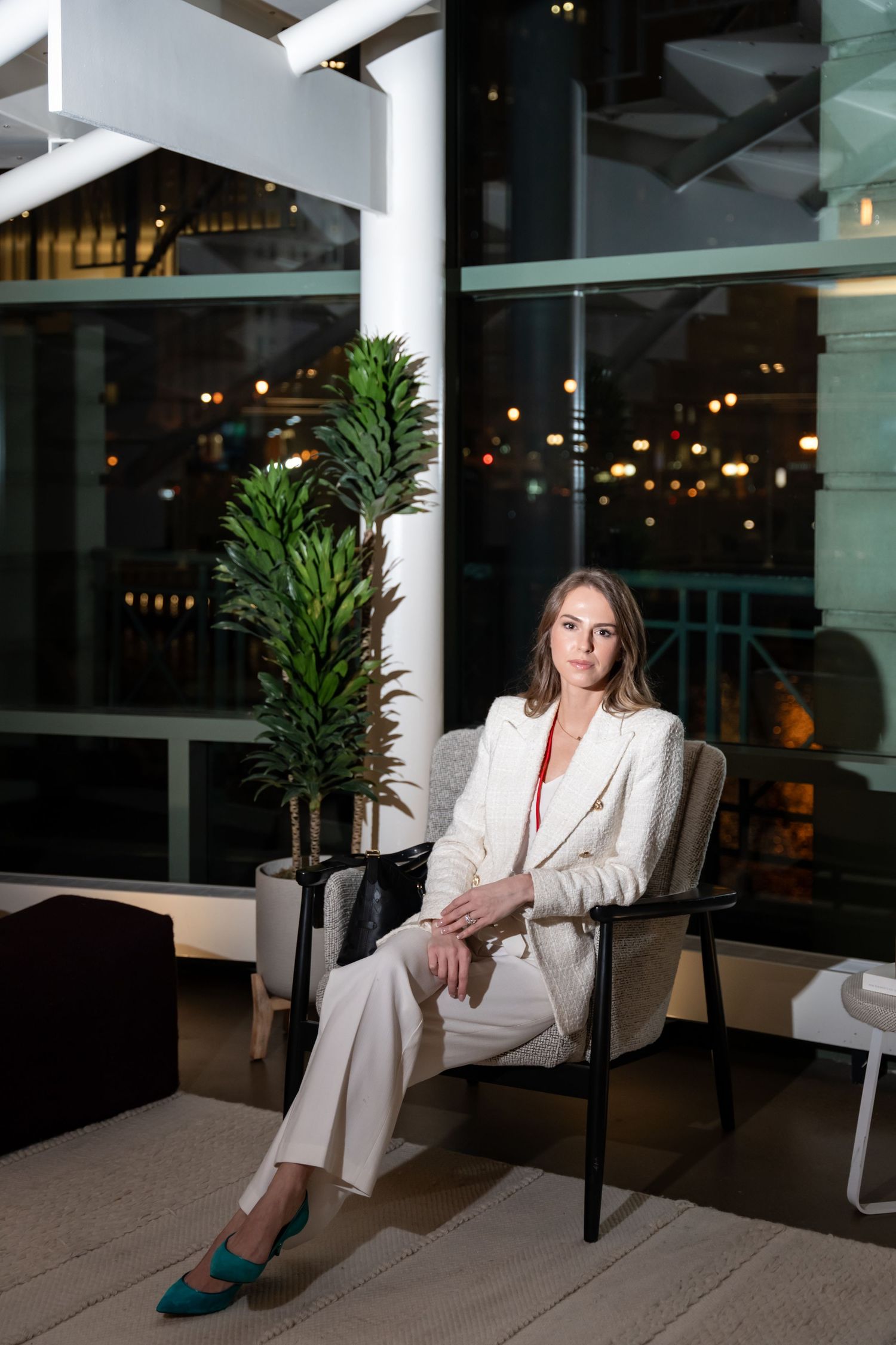 Woman in white suit sitting elegantly in modern space with glass windows and potted plants.