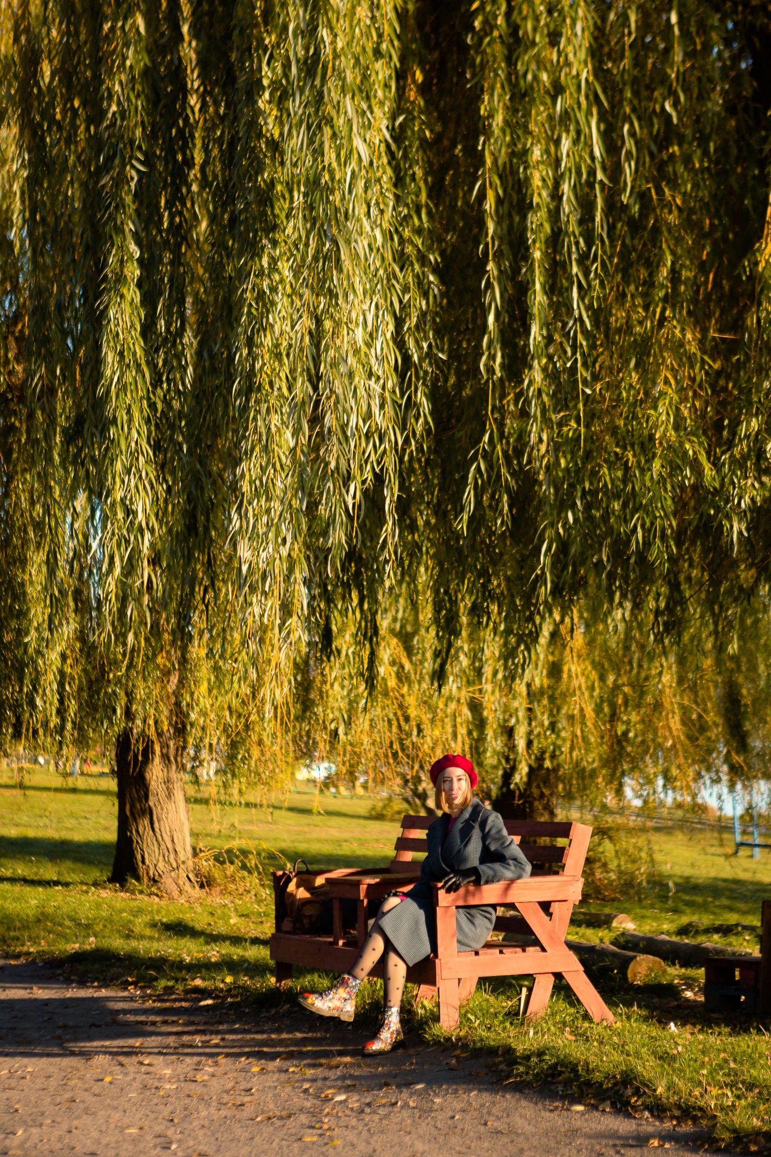 Person in dark coat sitting on wooden bench under weeping willow tree in autumn park.