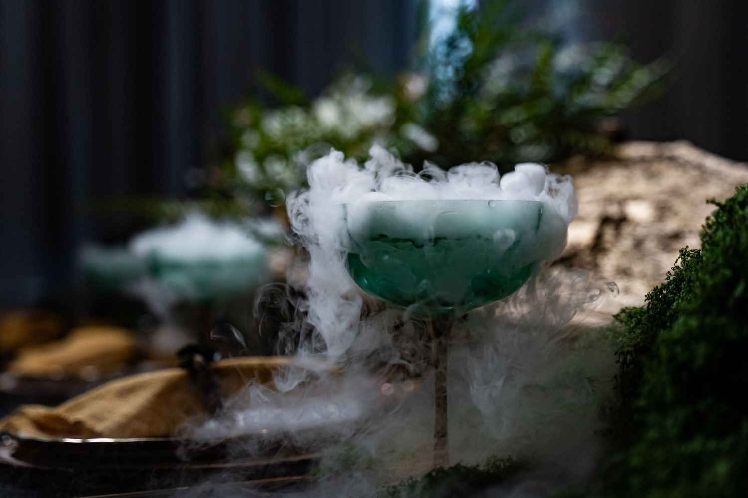 Smoking cocktail in green glassware creating dramatic fog effect against dark background.