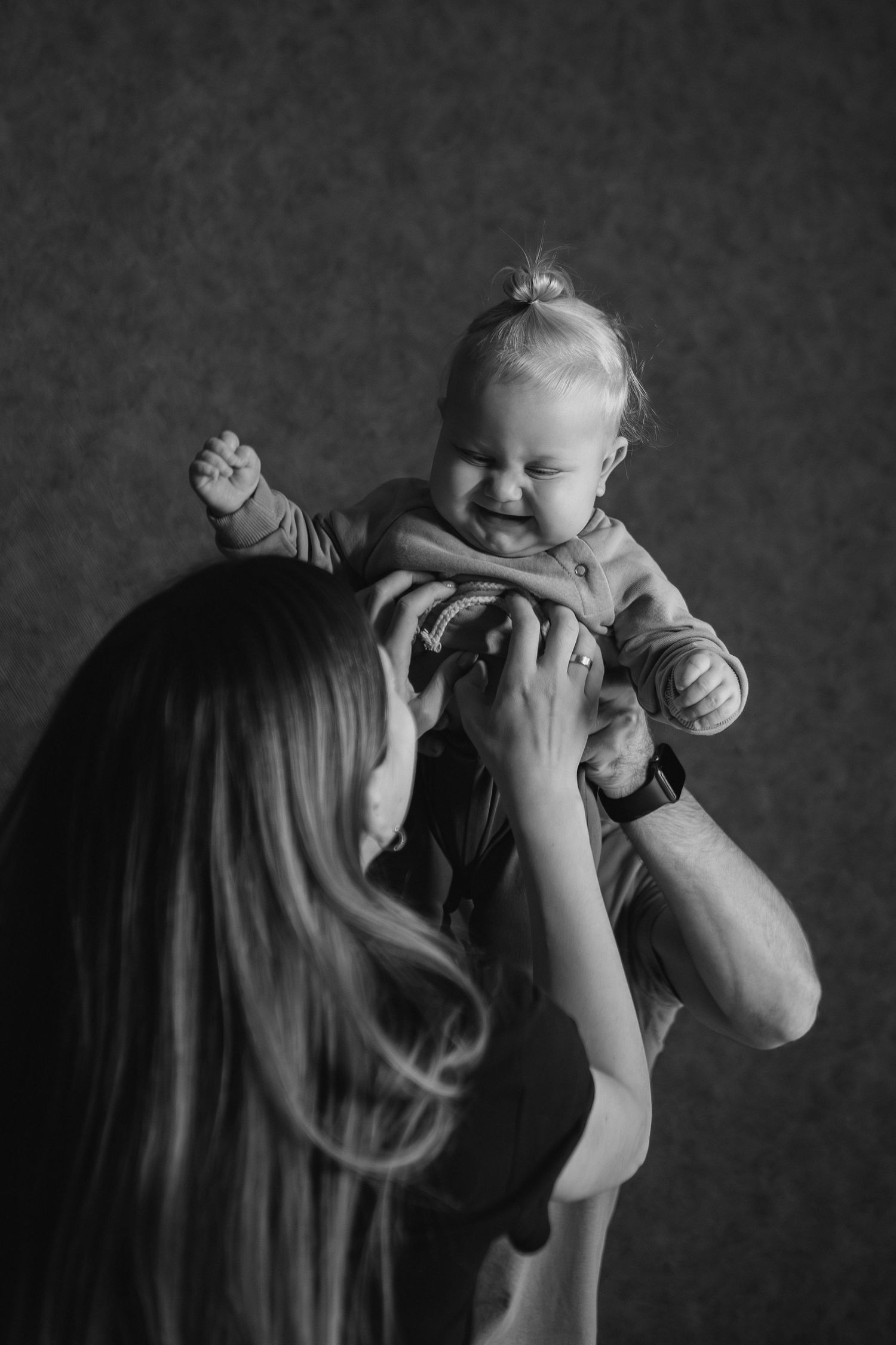 A black and white photo series showing a parent lifting and playing with a smiling baby against a dark background.
