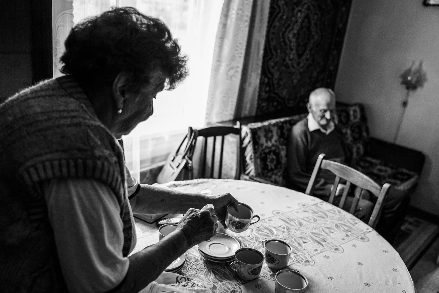 Two people sit at a table sharing tea and conversation in a dimly lit room.