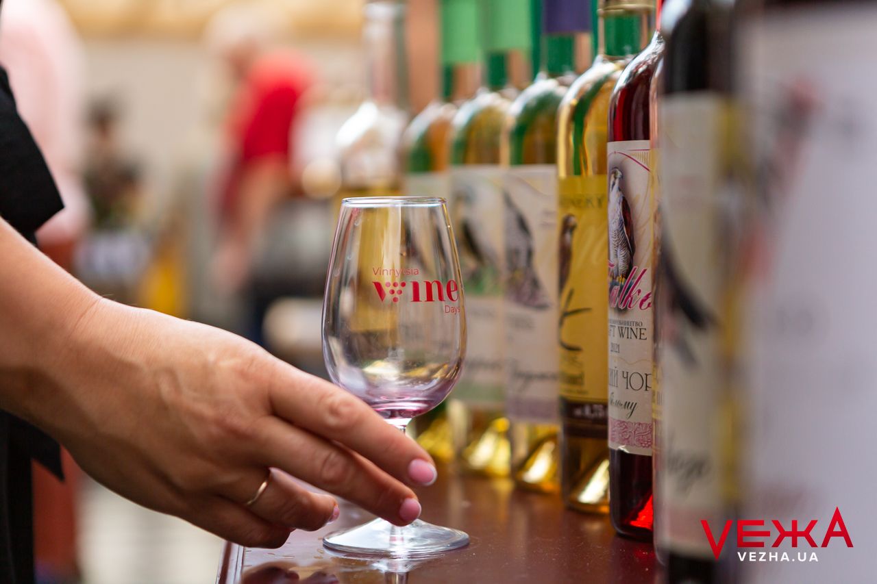 A hand reaches for a wine glass at a tasting event with multiple bottles lined up in the background.