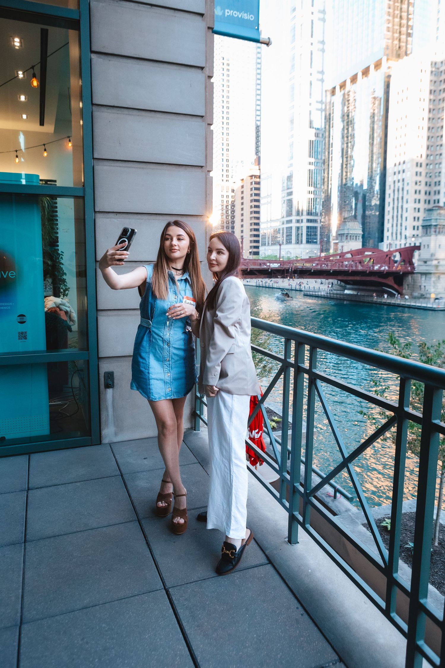Two friends taking selfies on a waterfront balcony with Chicago's riverwalk in the background.