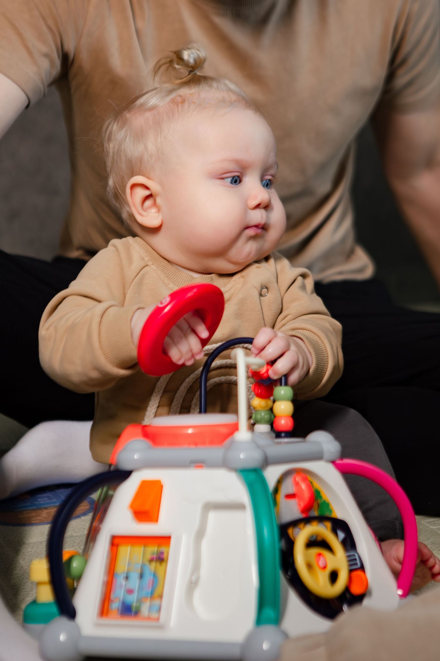 Baby in tan sweater explores colorful activity toy with handles and buttons from different angles.