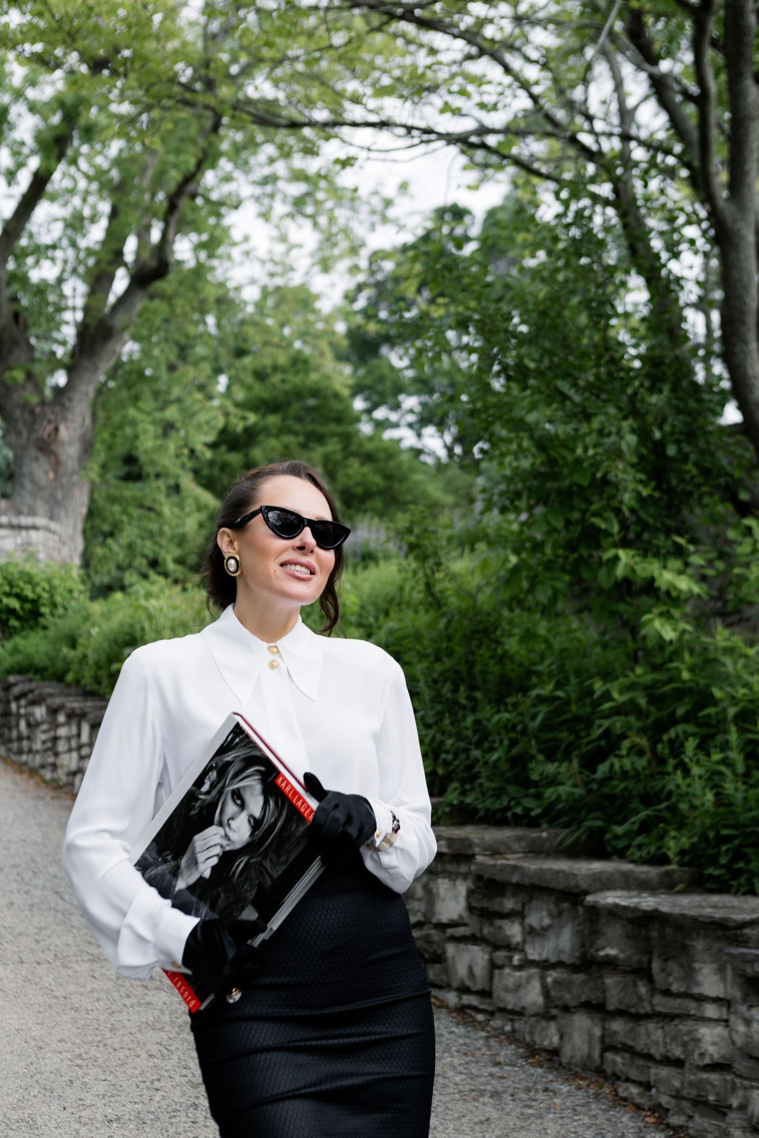 Woman in white blouse and sunglasses poses outdoors along stone wall path in spring garden setting.