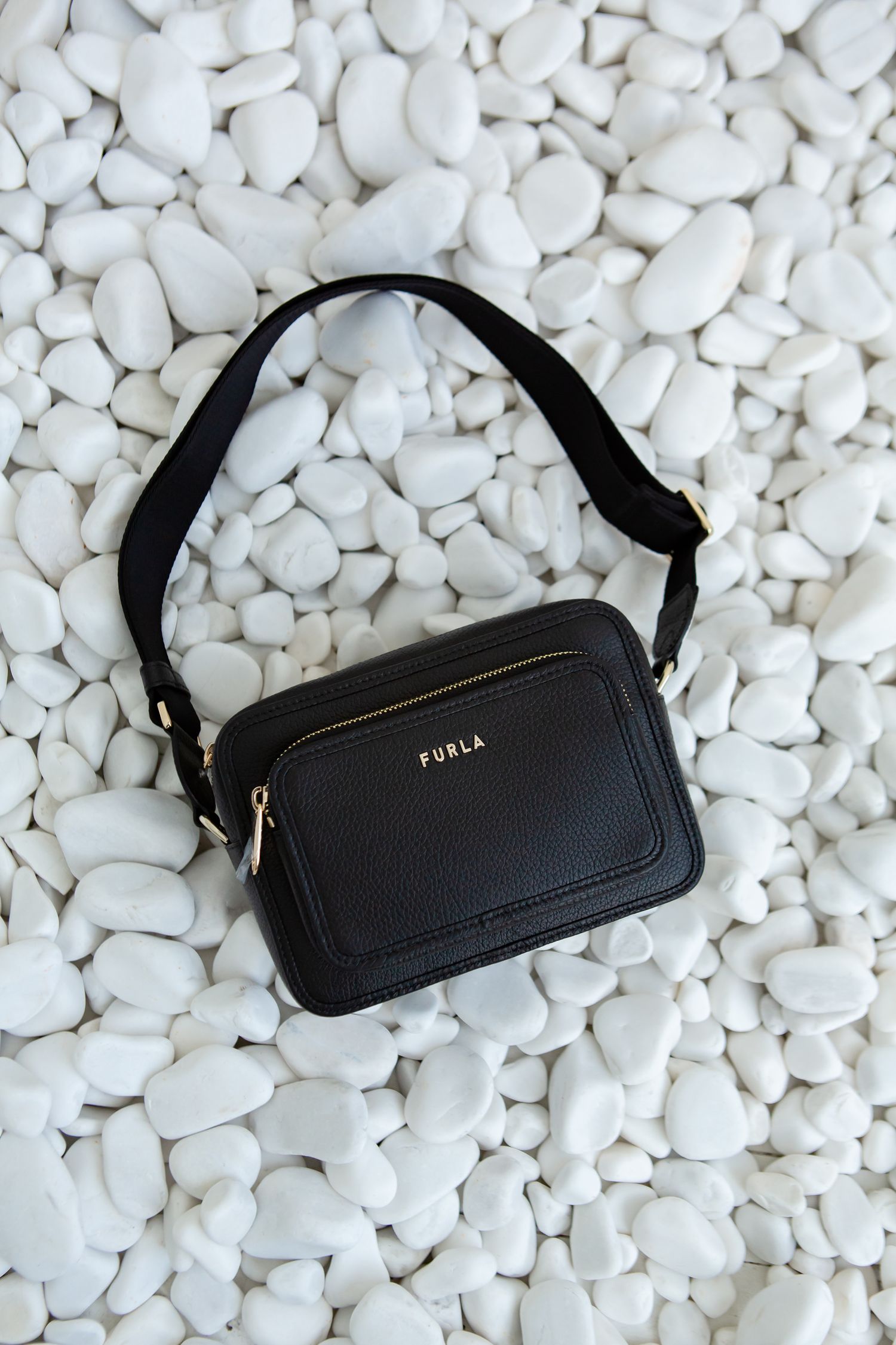 Black Furla crossbody bag displayed on white pebble background.