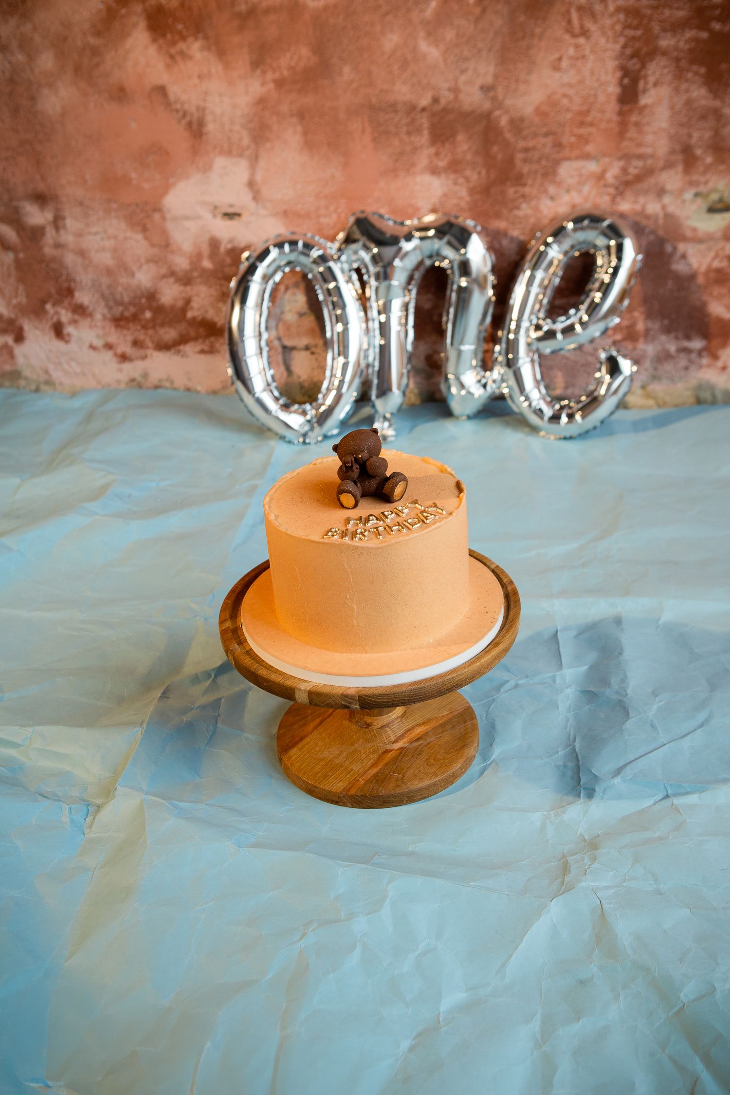 Orange birthday cake with silver 'ONE' balloon topper on wooden stand.