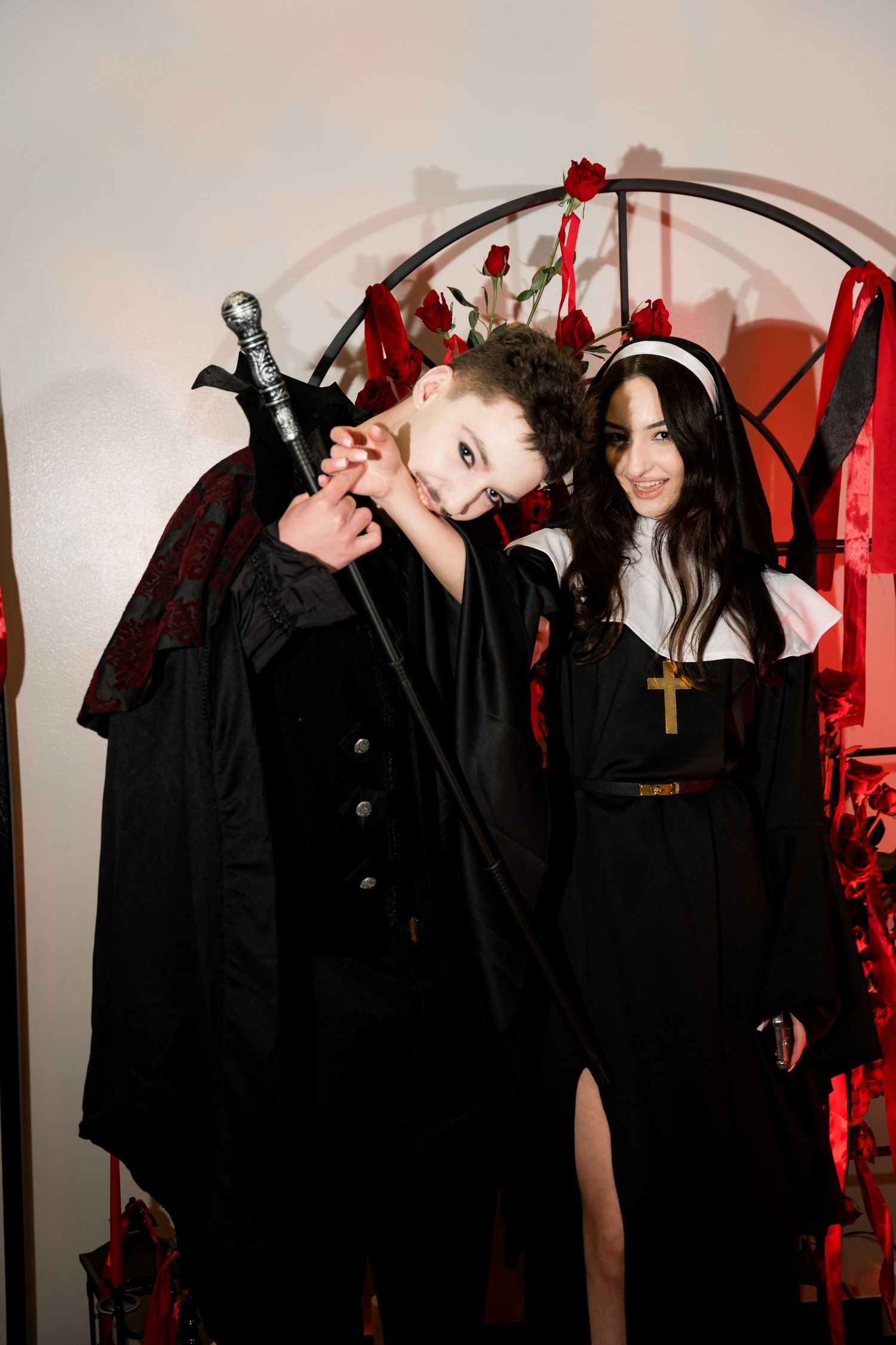 Two people in gothic Halloween costumes pose together in front of a red-lit backdrop with dramatic styling.