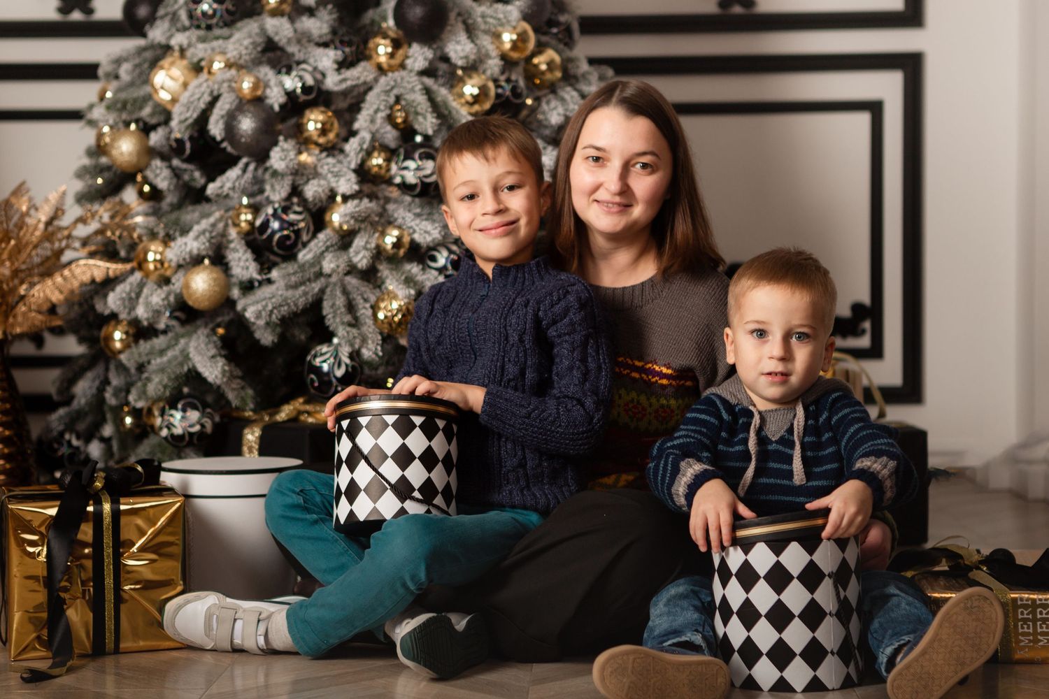 Siblings sitting by decorated Christmas tree with matching checkered gift boxes.
