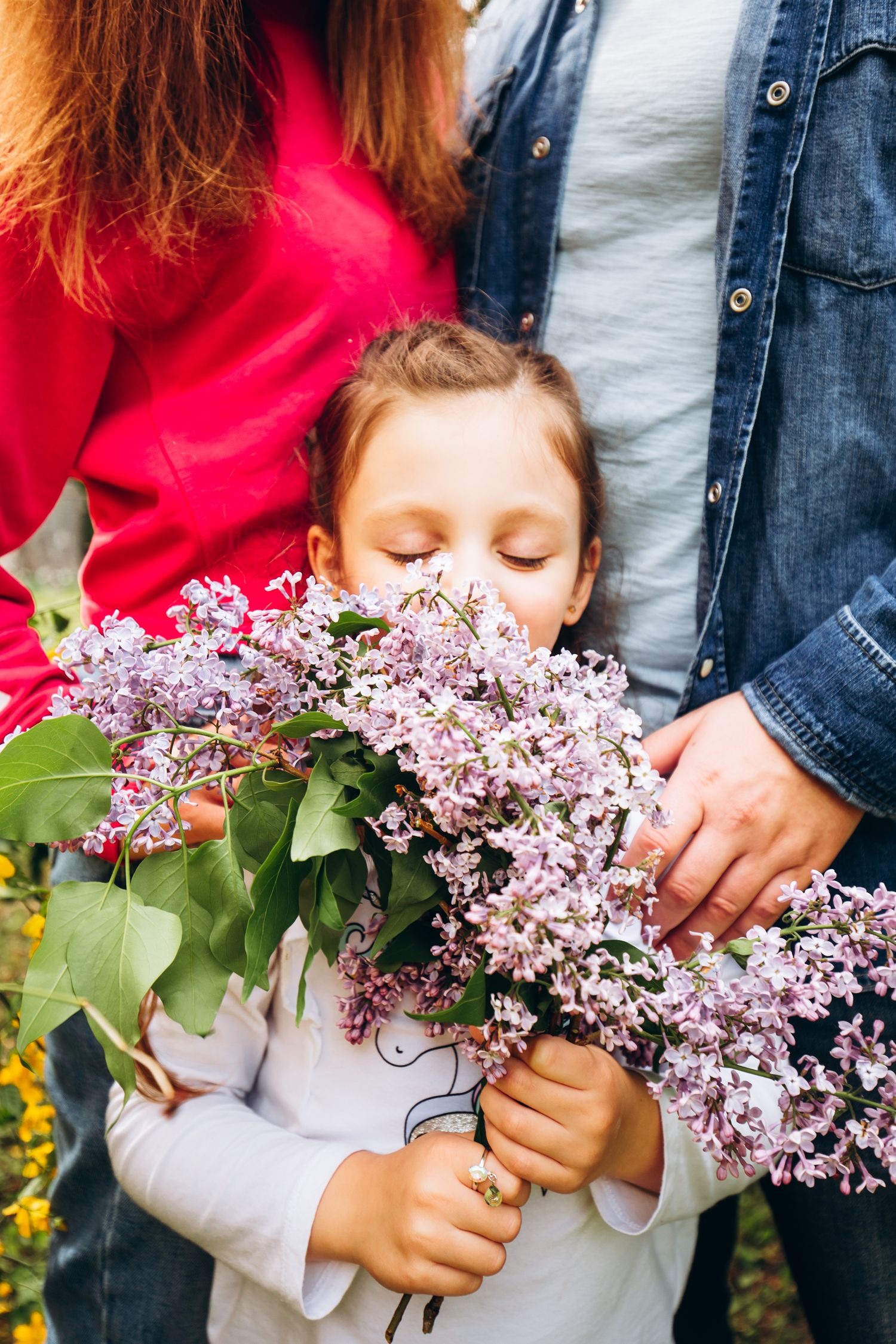 Child holding lilac flowers while embraced by parents in garden setting.