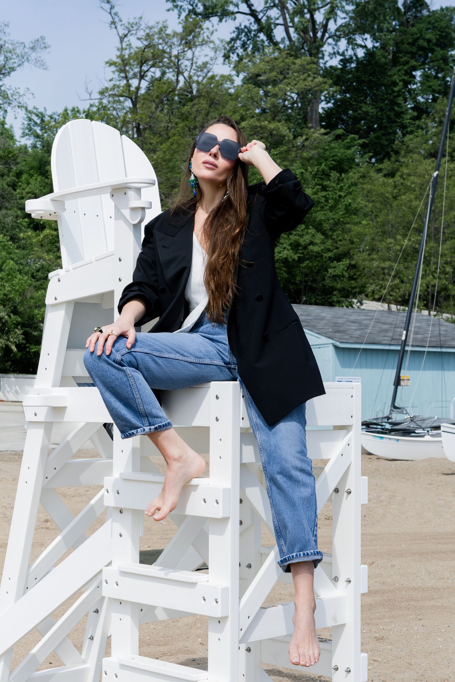 A woman in a black blazer and jeans sits casually on a white lifeguard chair near a pool and sailboats.