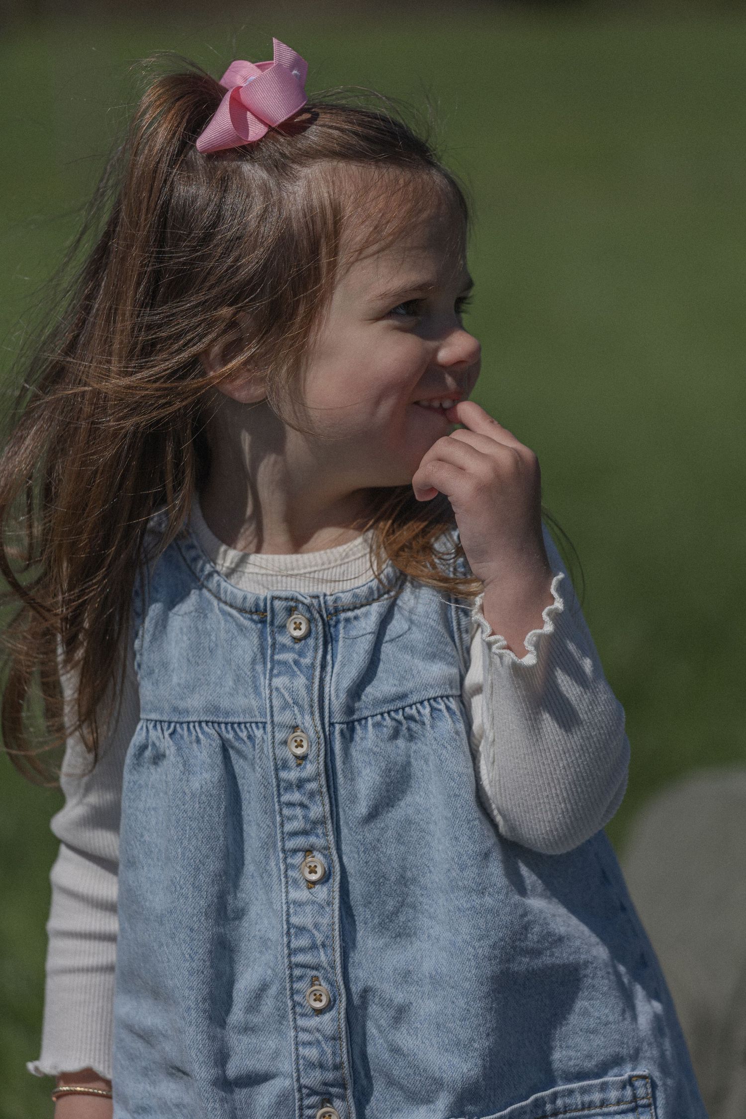Young girl in denim dress with pink bow smiling outdoors in sunlight.