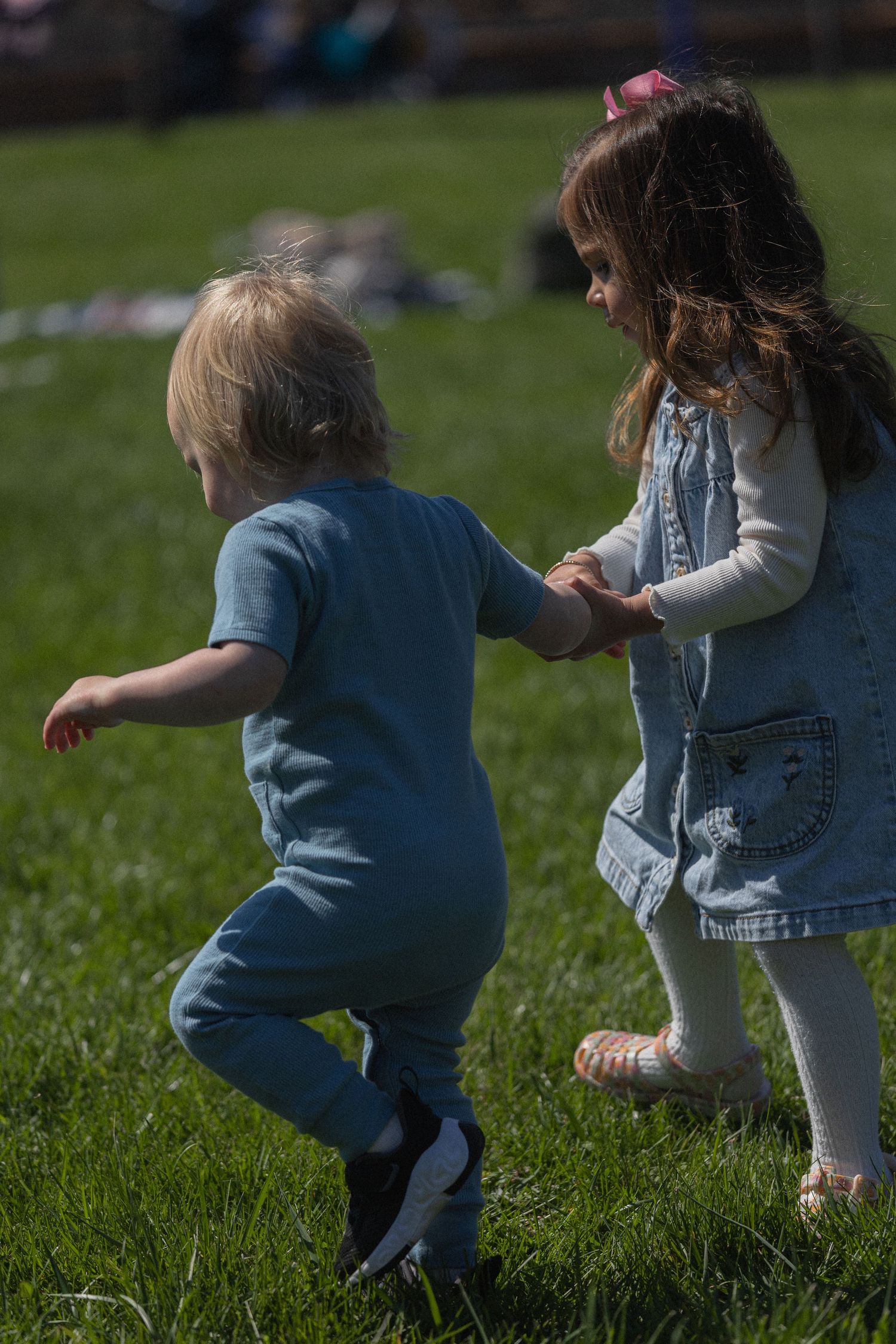Two young children holding hands and playing on green grass outdoors on a sunny day.
