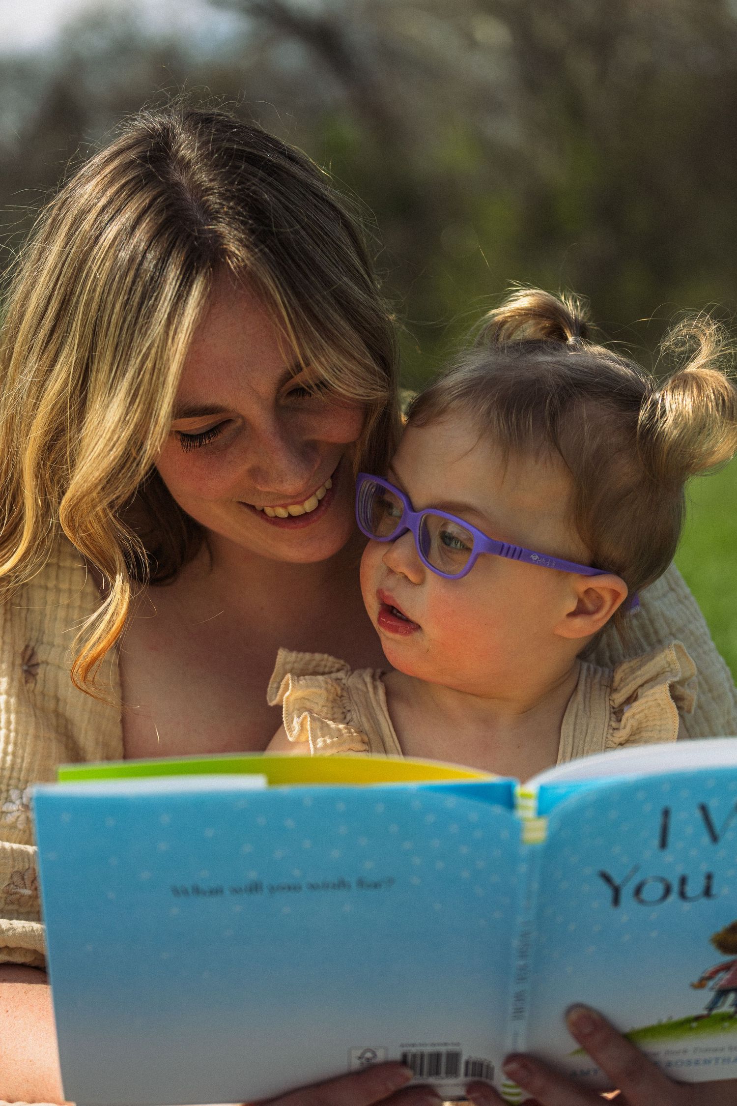 A smiling mother reads a colorful picture book with her toddler daughter wearing purple glasses outdoors.