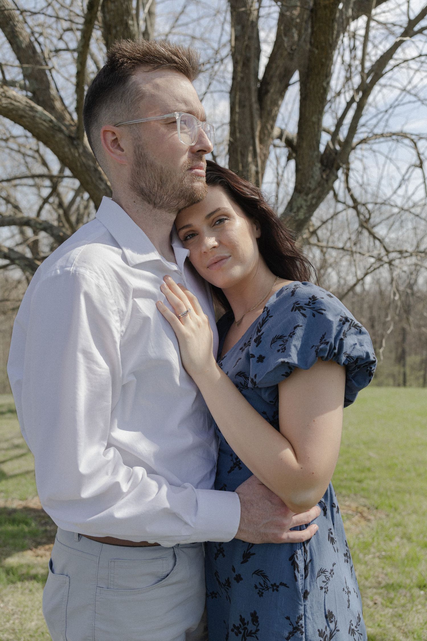 A couple embraces outdoors near bare trees, woman in blue floral dress leans on man in white shirt on a sunny day.