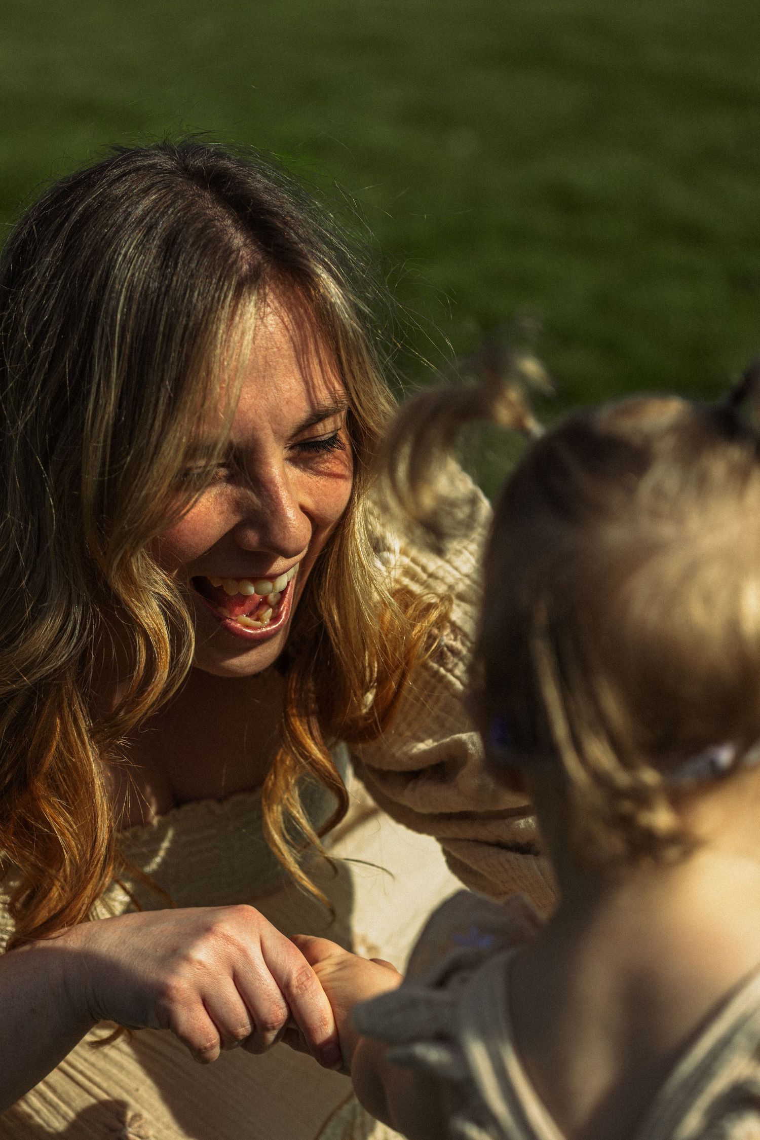 A laughing woman enjoys a sunny outdoor moment with a young child on green grass.