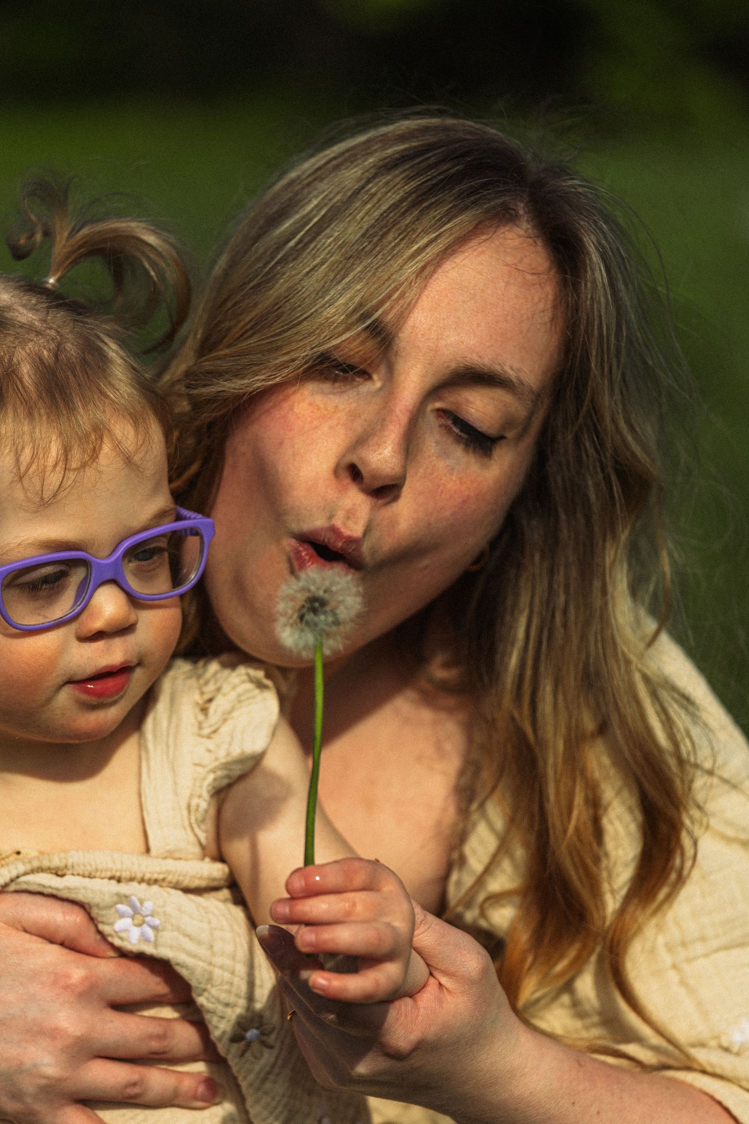A mother with freckles blows a dandelion wish with her toddler daughter wearing purple glasses outdoors.