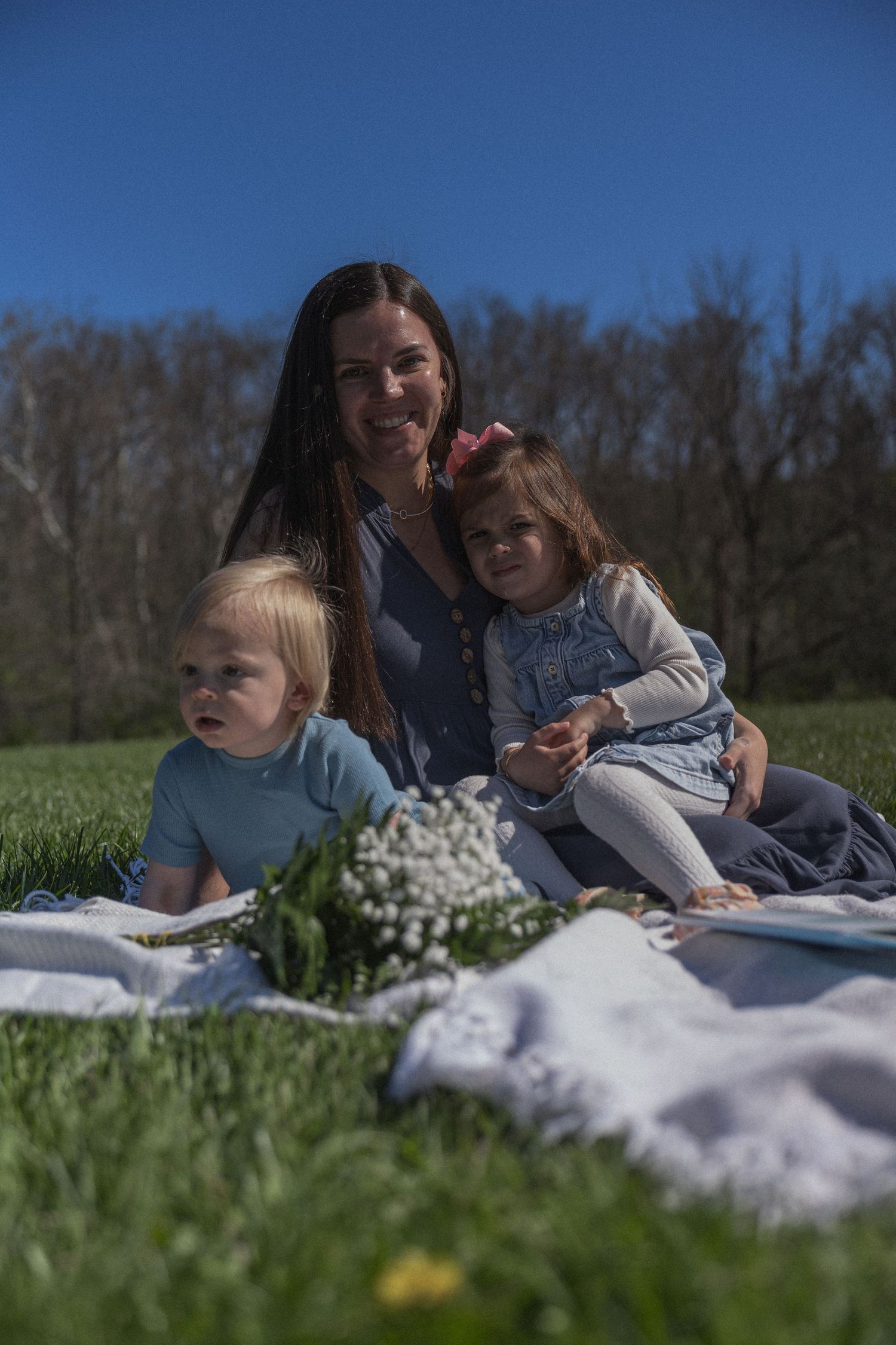 A smiling mother sits on a white blanket in a sunny park with her two young children and a bouquet of white flowers.