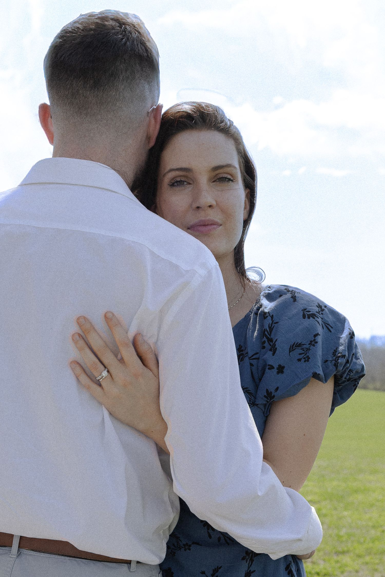 A woman in a blue floral dress embraces a man in a white shirt outdoors, showing an engagement ring on her hand.