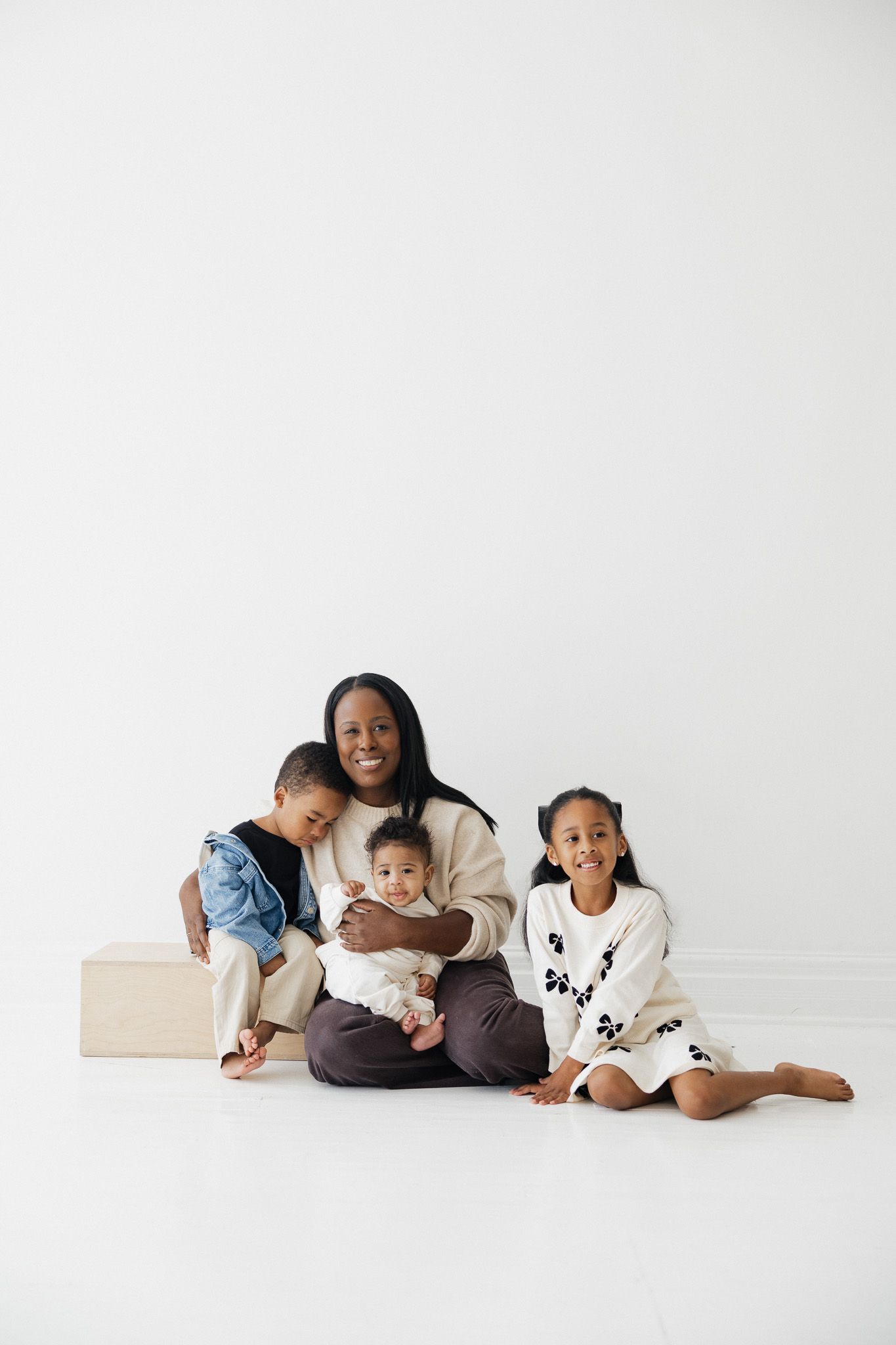 A mother sits on the floor smiling with her three young children against a white background.