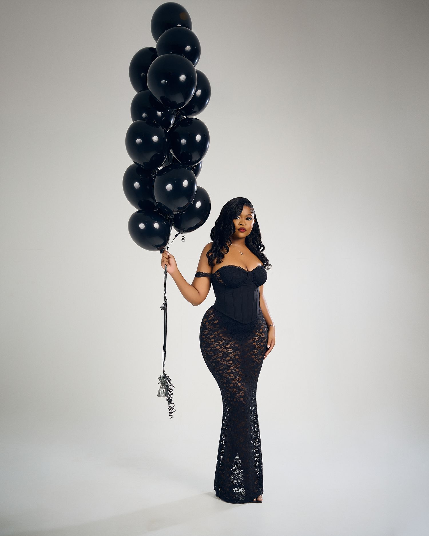 Fashion model in black lace gown poses with cluster of black balloons against white background.