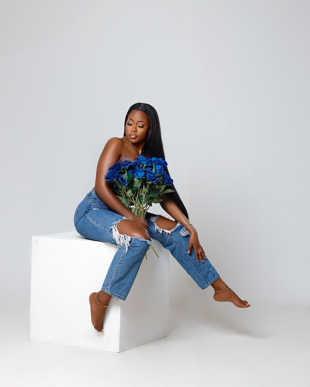 Fashion model poses on white cube wearing distressed denim jeans and holding blue flowers in artistic photoshoot.