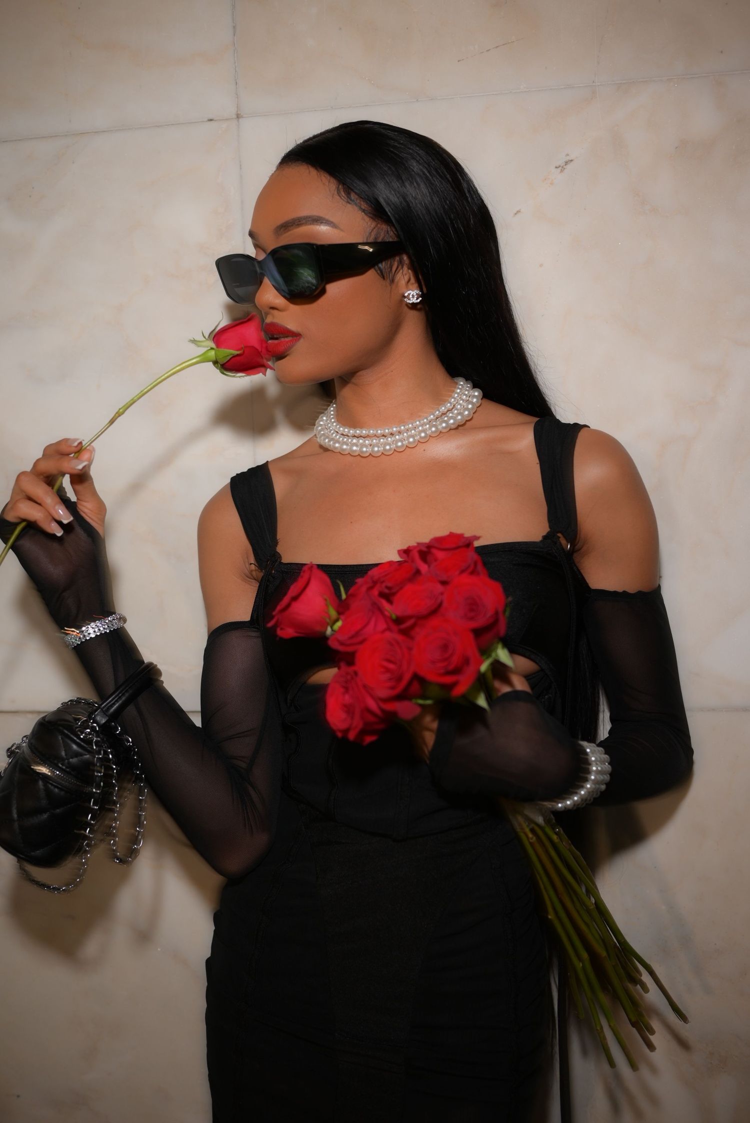 Elegant pose with red roses, black dress, and pearl necklace against light wall.