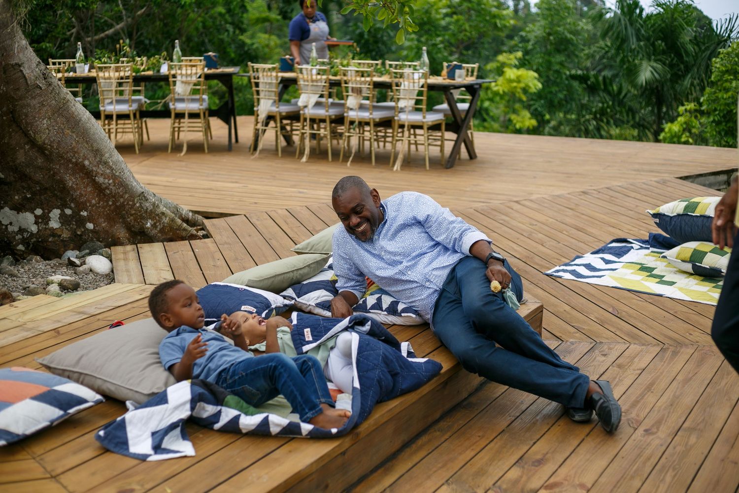 A man and two children relax on a wooden deck with colorful blankets, smiling outdoors near a dining area.