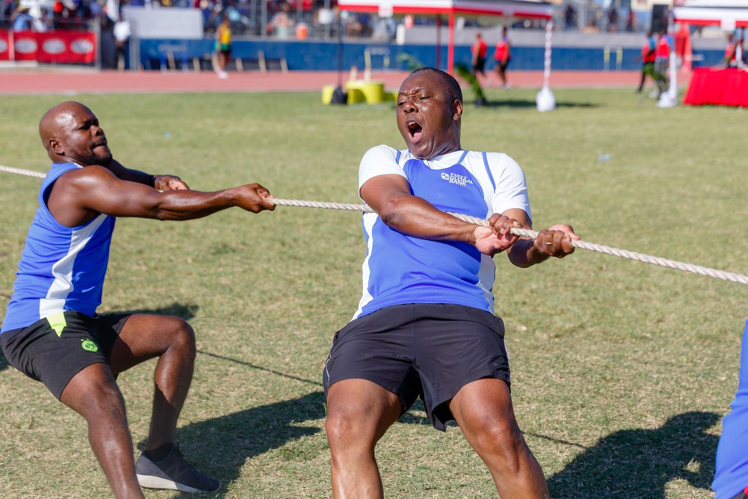 Two men competing in a tug of war event at an outdoor sports stadium, straining with effort on a grassy field.