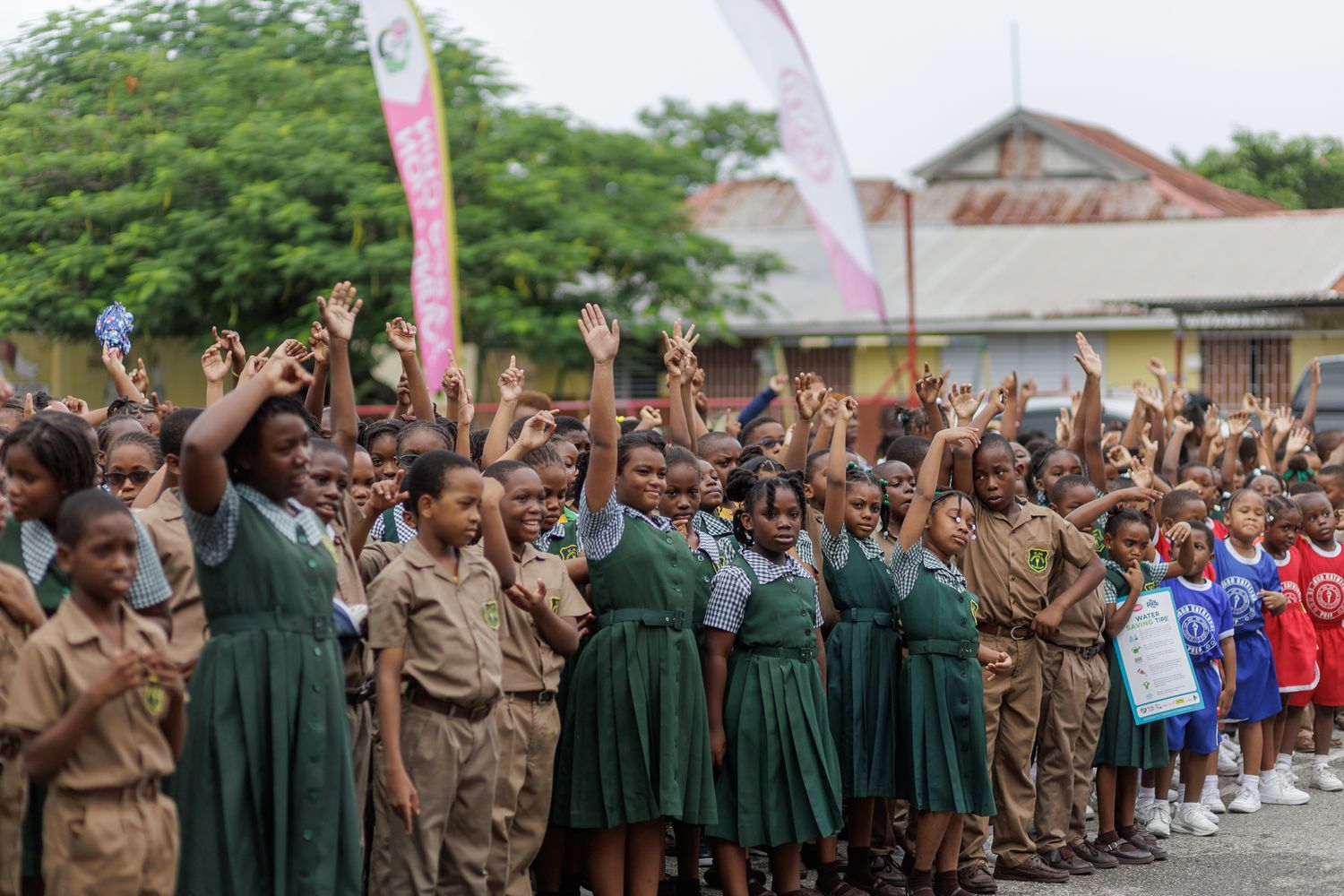 Schoolchildren in uniforms raising hands at an outdoor gathering with pink banners in an African community event.