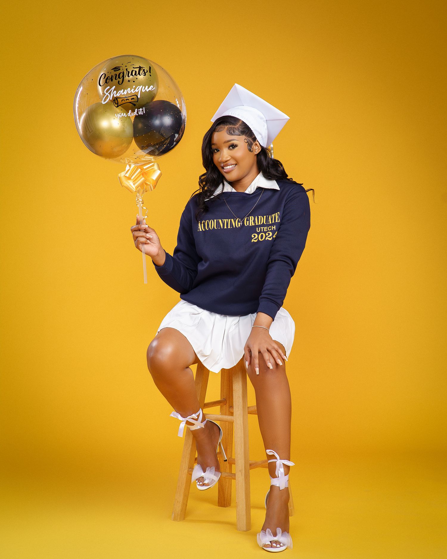 Graduate in navy sweatshirt and white skirt sits on wooden stool holding celebration balloons.