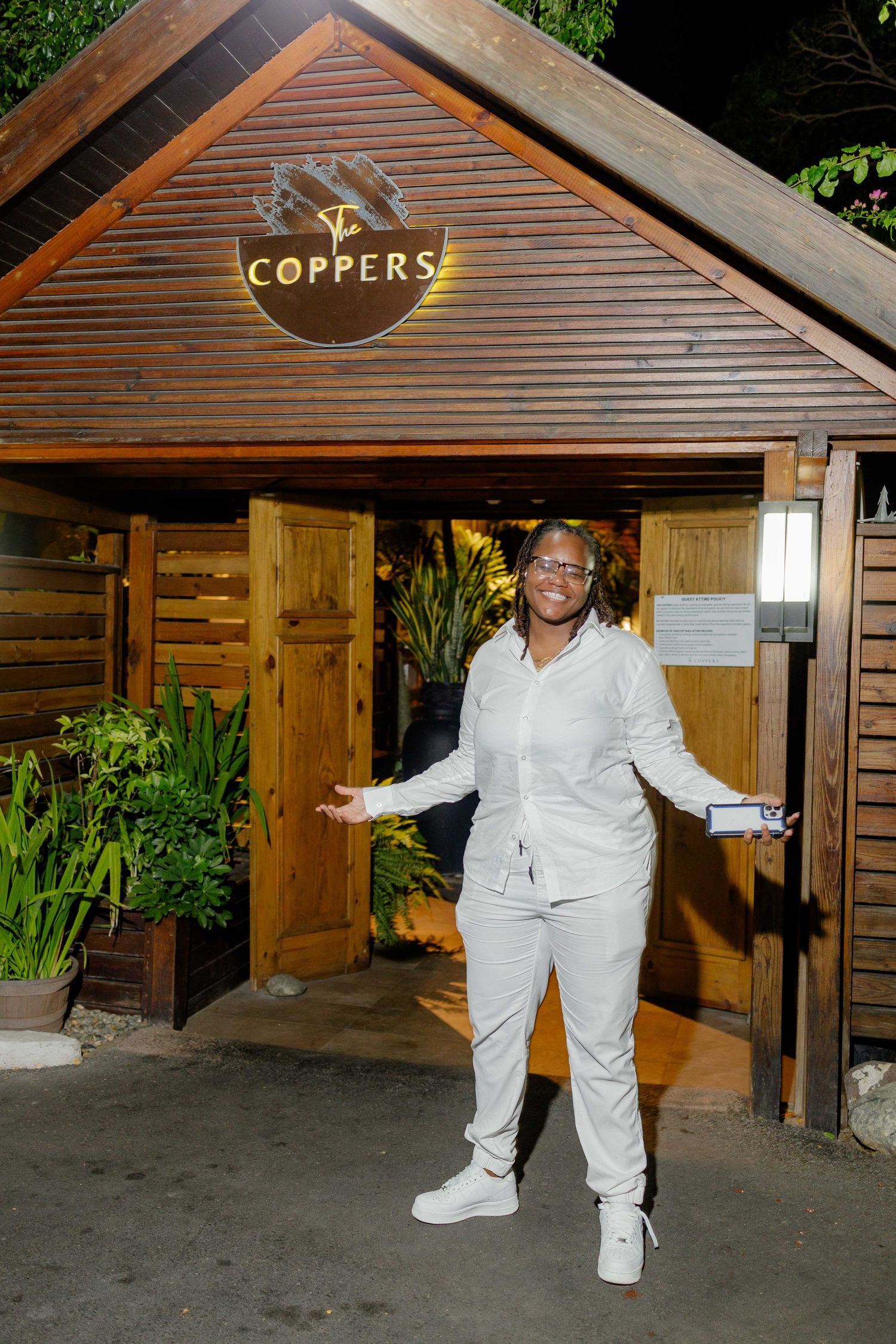 Person in all-white outfit posing under wooden structure with Coppers signage.